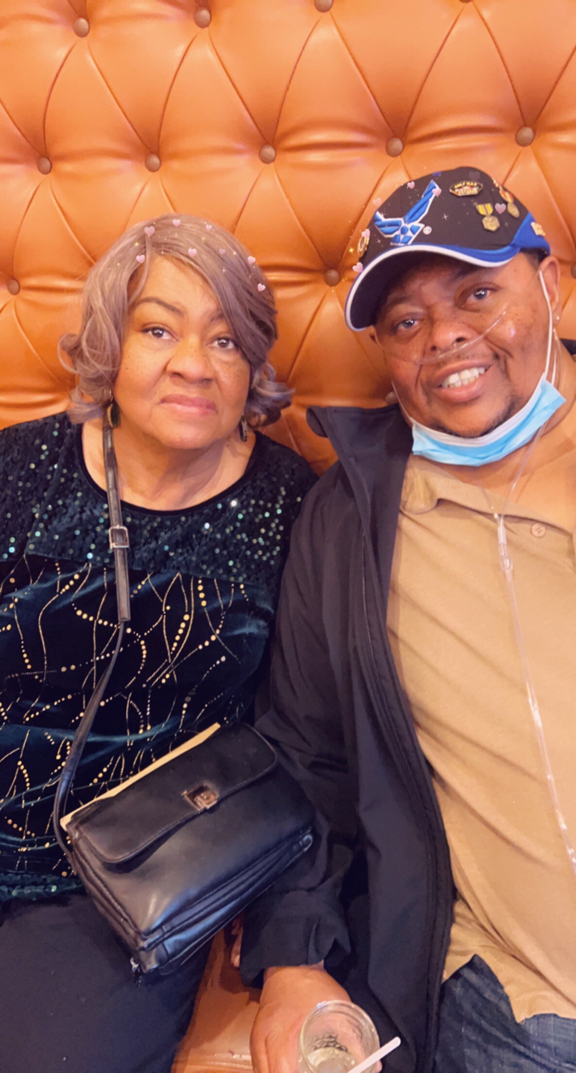 Two smiling adults sit close together on an orange tufted bench; woman in a black sequined dress on the left, man wearing a cap and mask pulled down on the right, holding a drink.