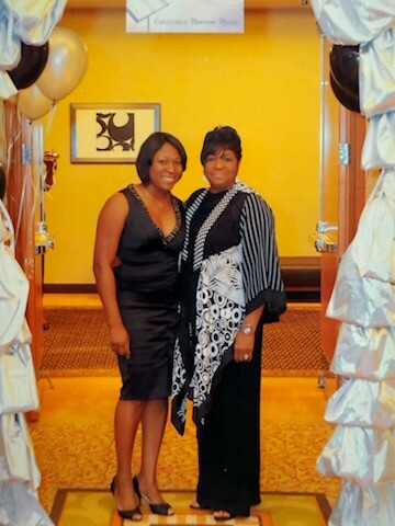Two women in black outfits pose together in a decorated doorway at an indoor event, with white drapery, balloons, and a yellow wall behind them.
