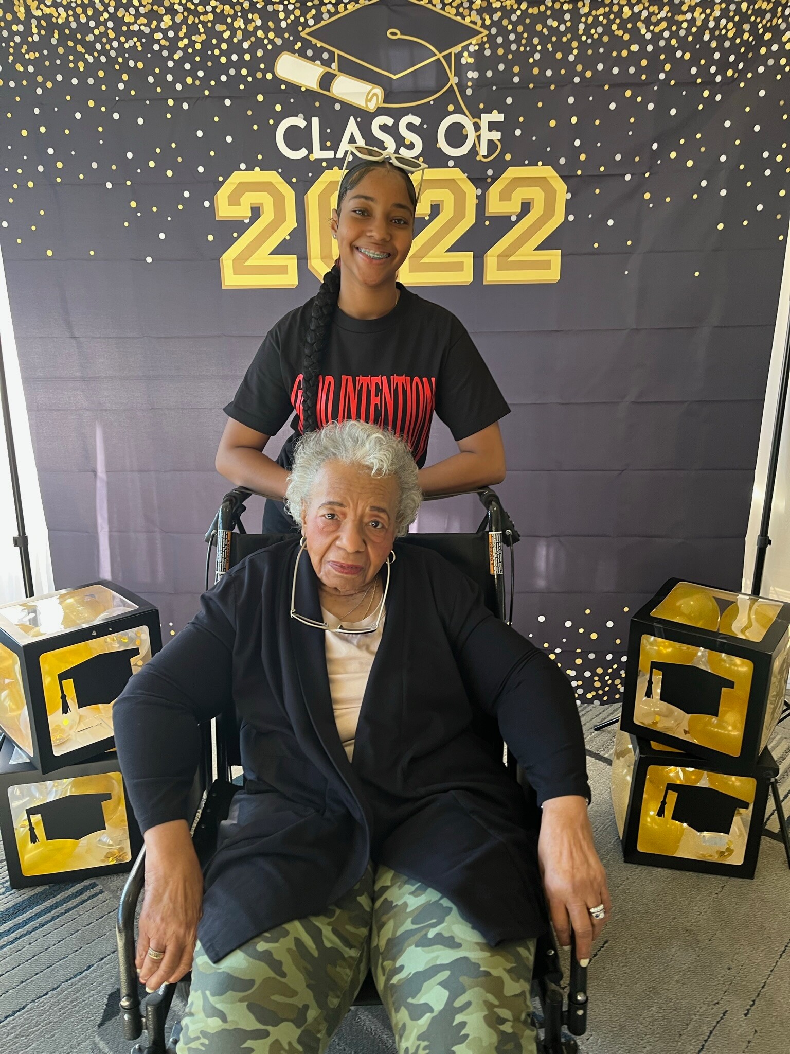 Young woman in a black shirt behind an elderly woman in a wheelchair, posing in front of a Class of 2022 backdrop.