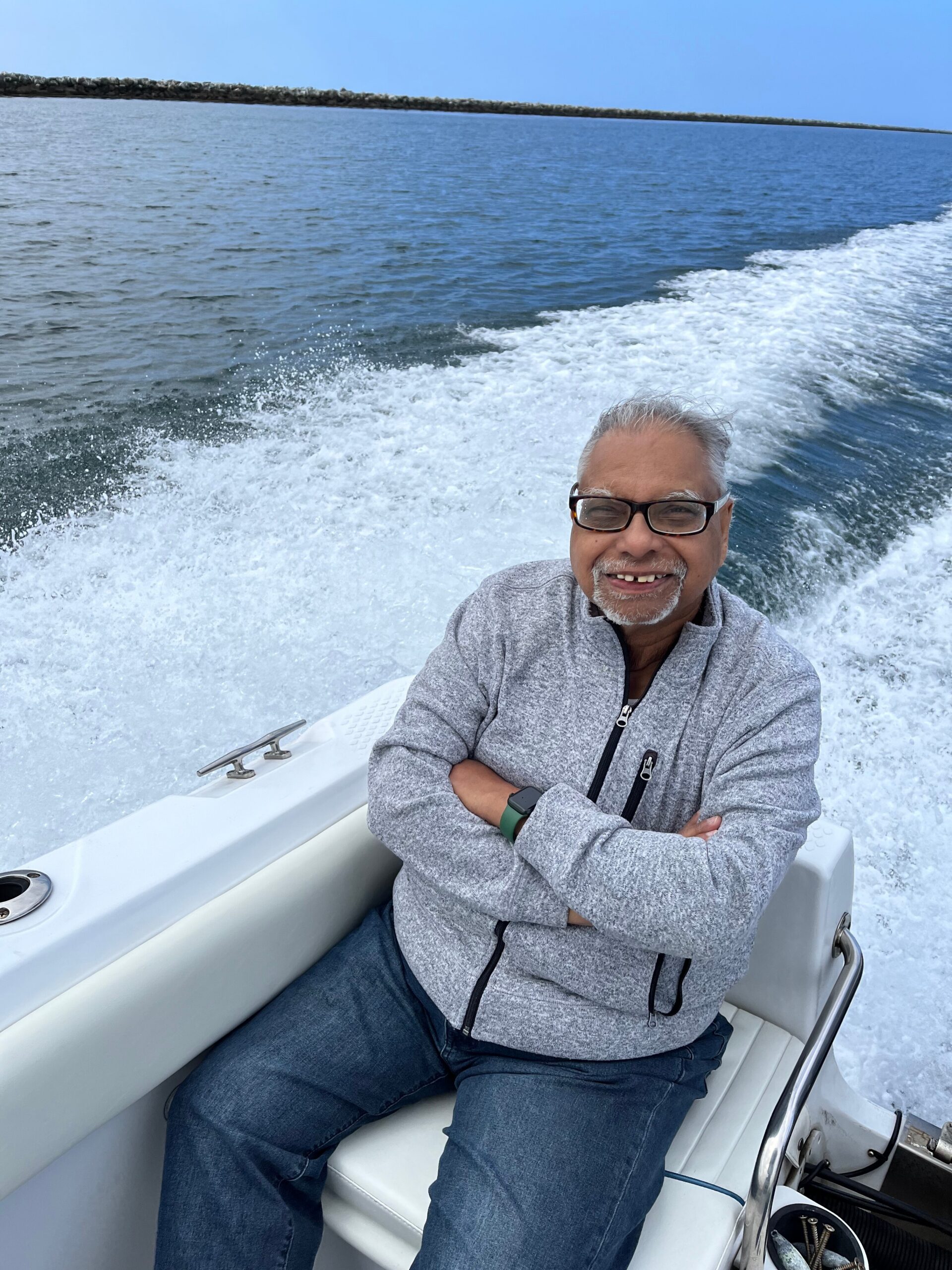 Smiling man wearing glasses and a gray zip jacket sits on a boat, with ocean and wake behind him
