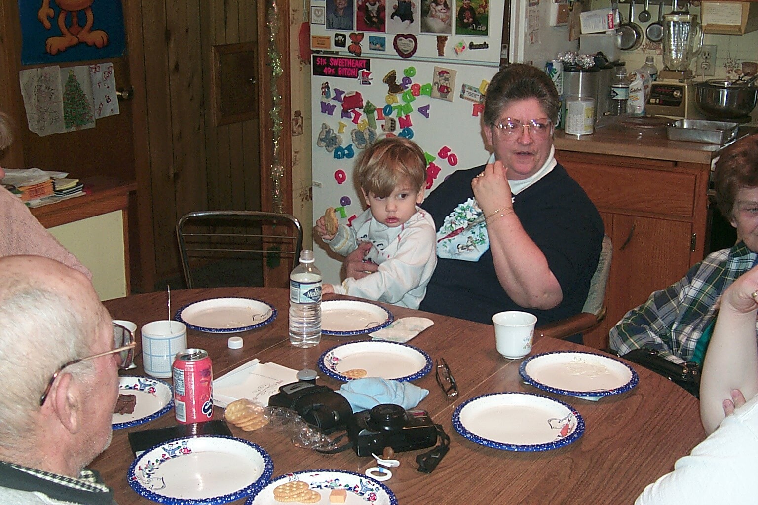 Older woman holding a small child at a round dining table with plates and snacks; a cluttered kitchen in the background.
