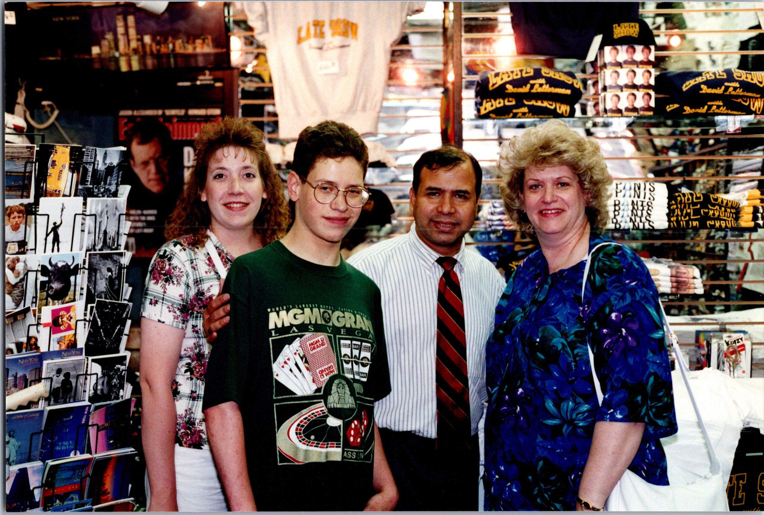 Four people pose and smile at the camera inside a souvenir shop with postcards and t-shirts on display behind themCon text: group photo in a store