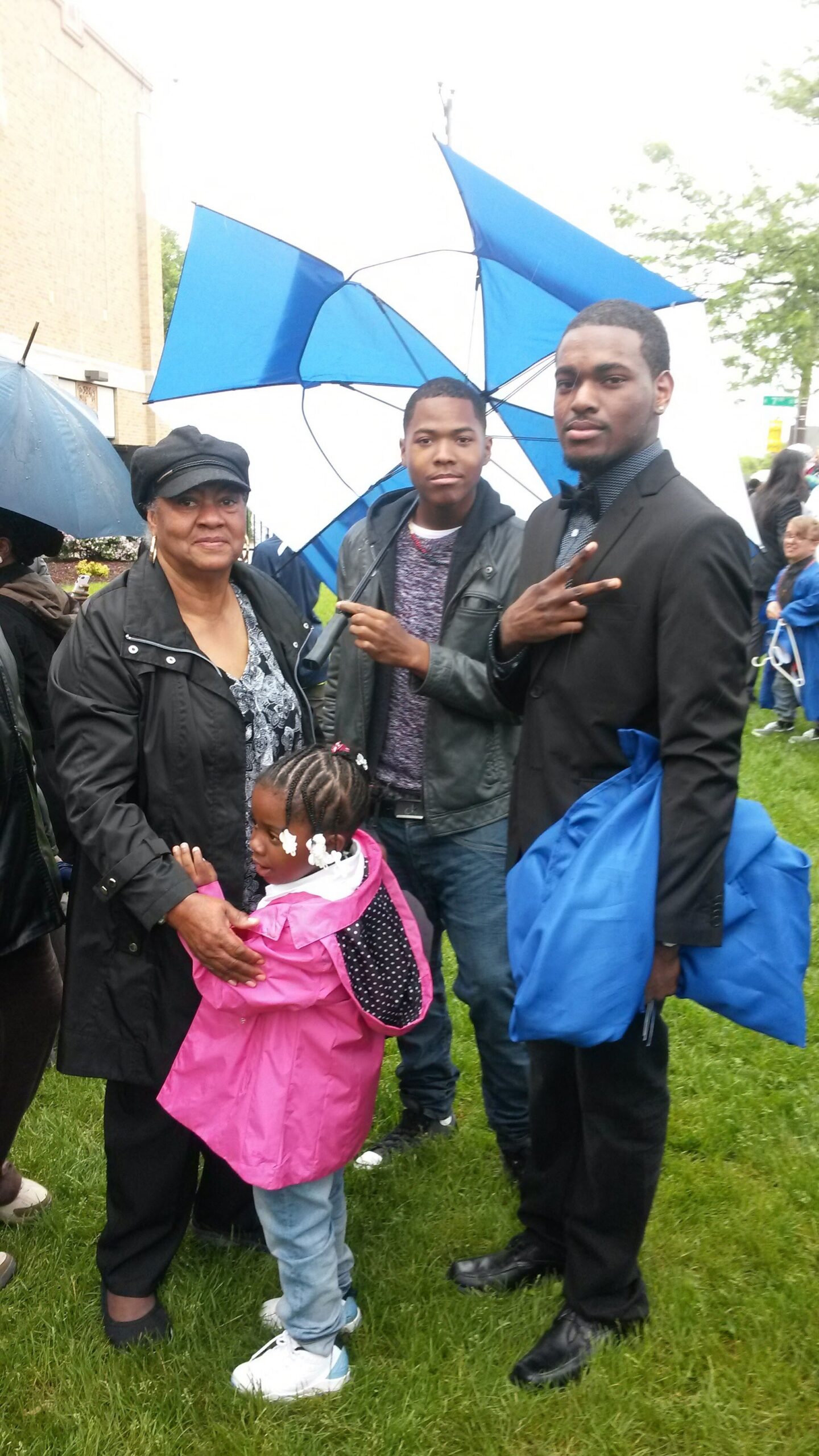 Group of people at an outdoor event holding blue umbrellas; a little girl in a pink raincoat stands with an older woman while two men pose nearby.