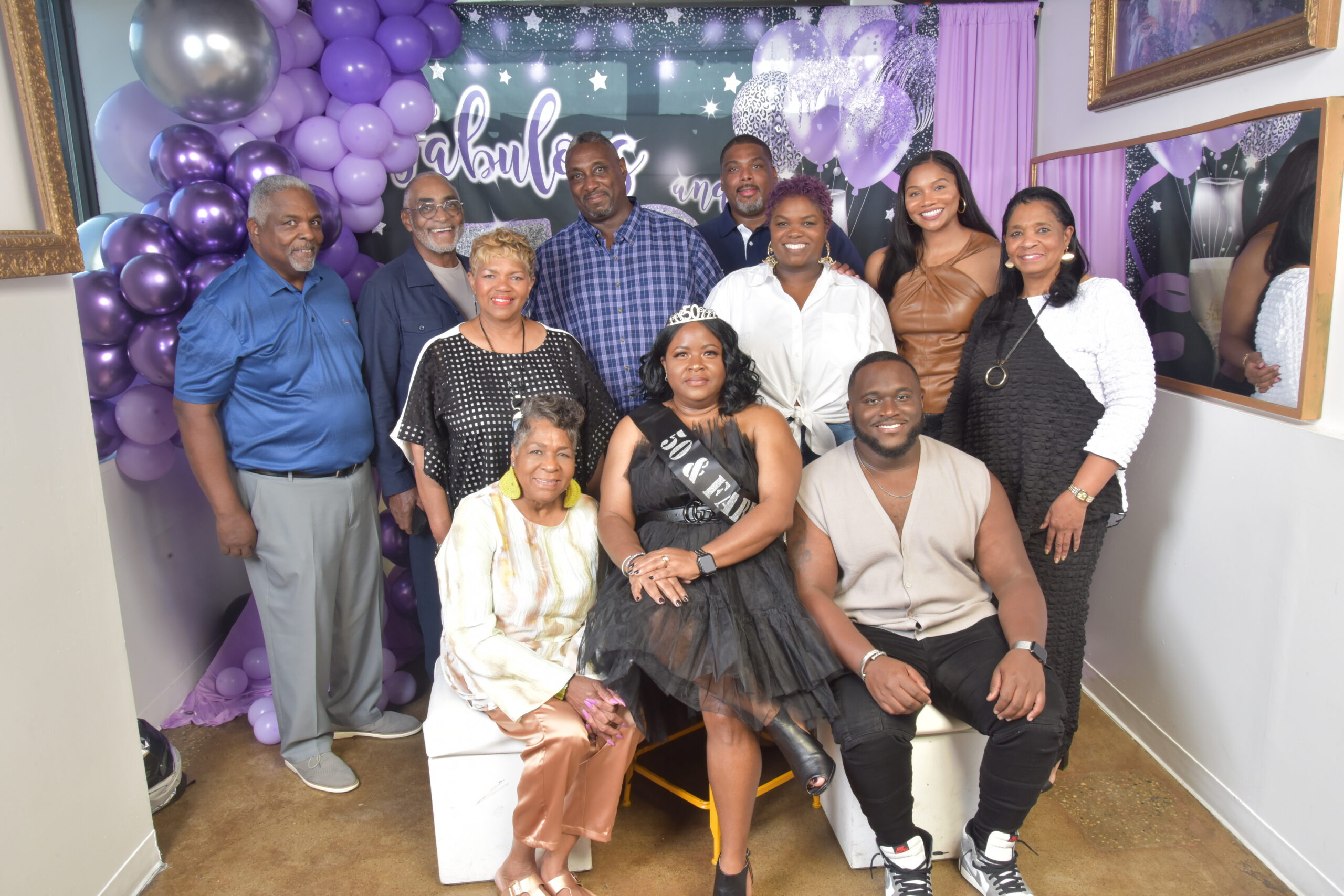 Group of adults posing together at a purple-themed celebration with balloons and a decorative backdrop behind them, indoors