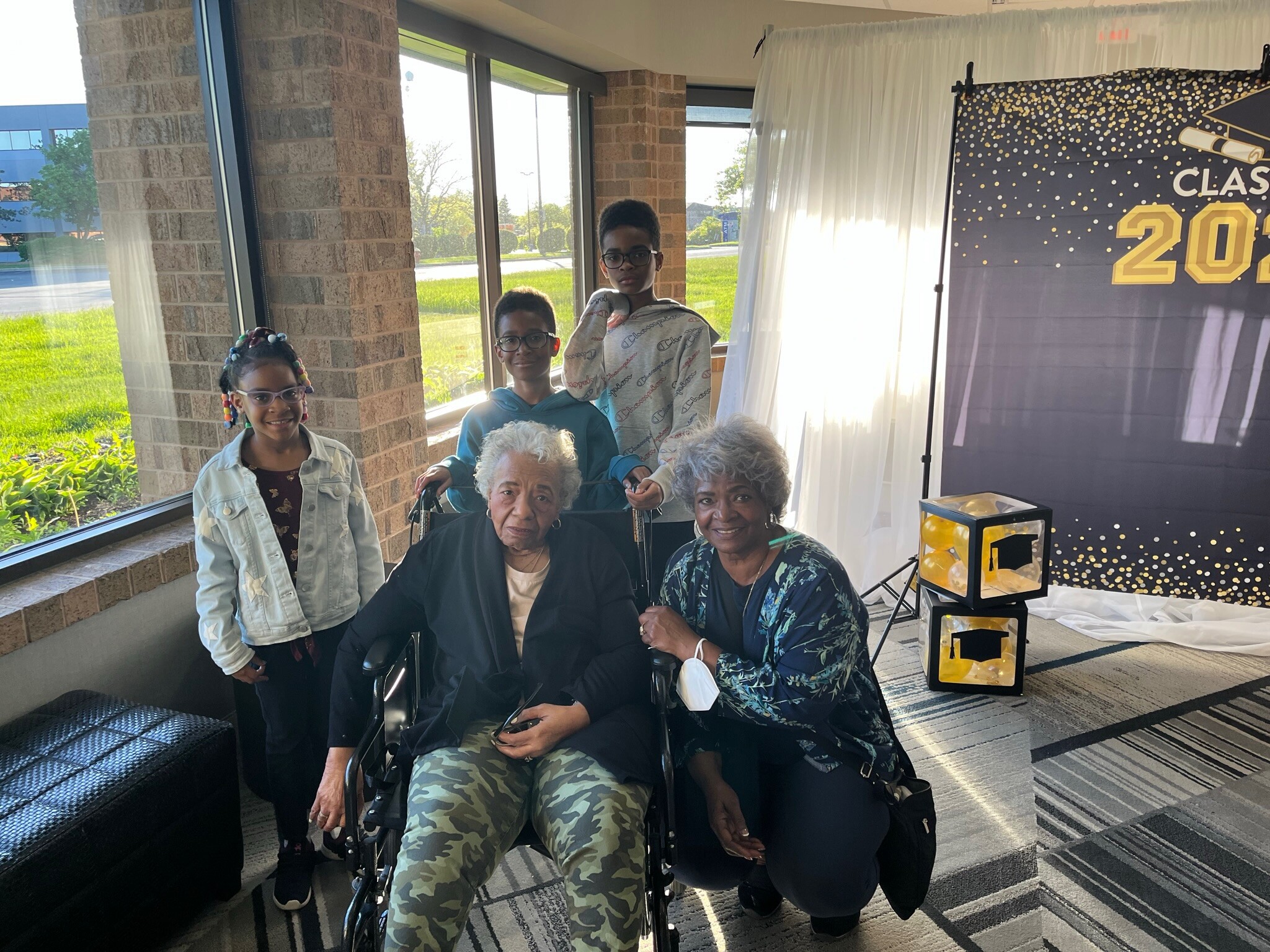 Group of children and seniors in wheelchairs posing indoors at a celebration, with a 'Class of 20' backdrop on the right and large windows behind.