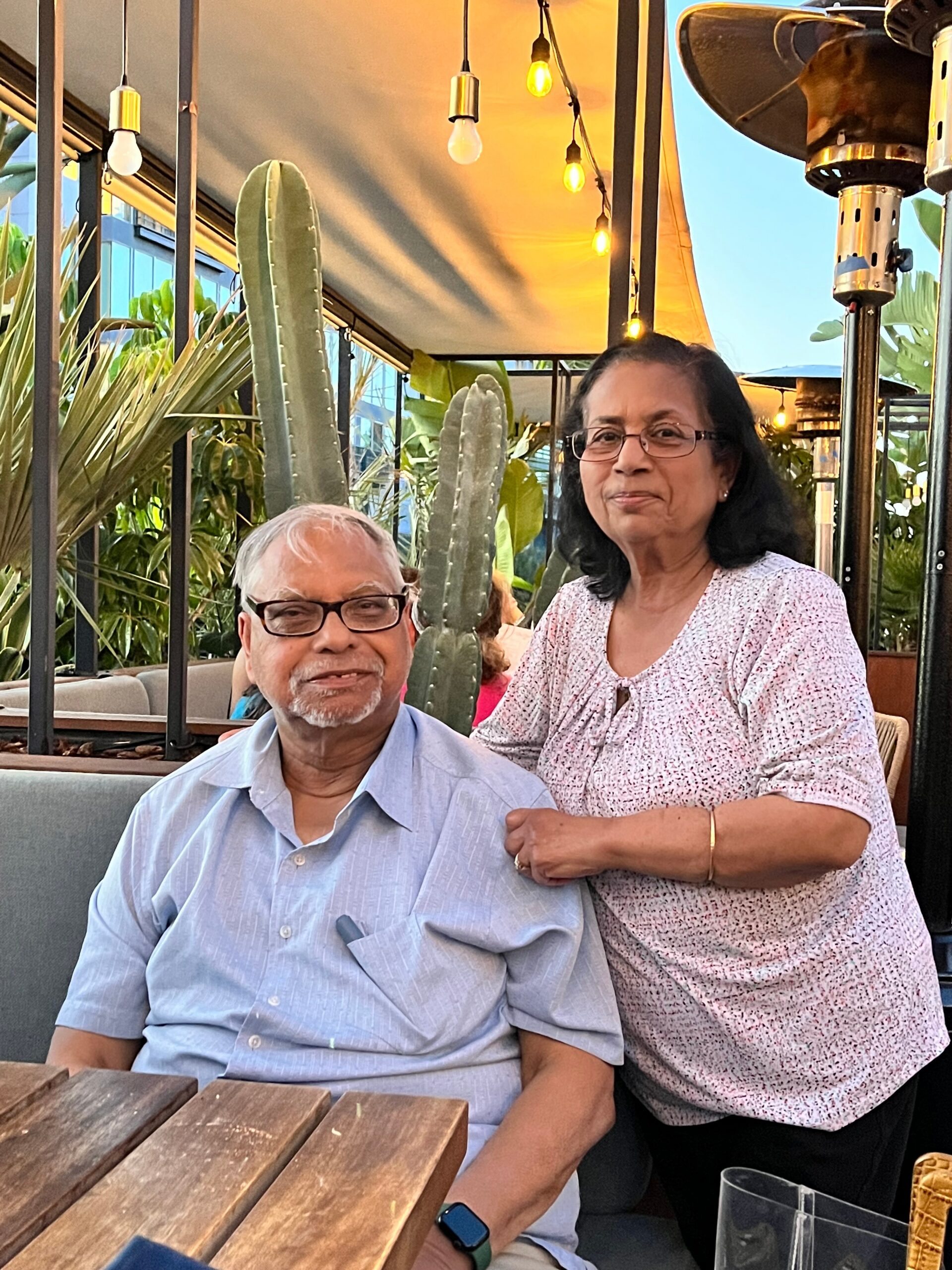 Smiling man in a light blue shirt sits at a wooden table, with a woman standing beside him and her arm around his shoulder, on an outdoor patio with string lights and cactus plants in the background.
