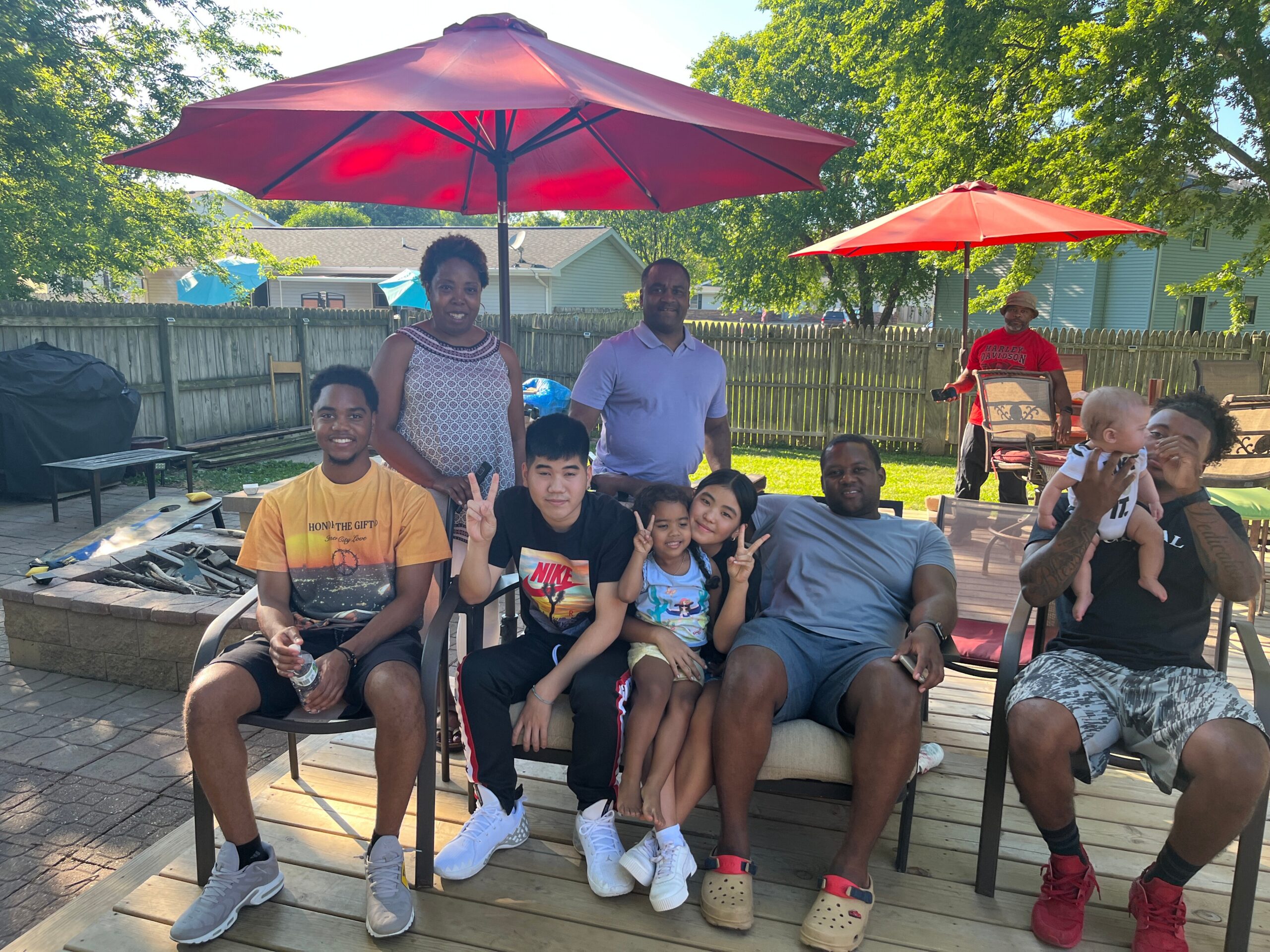 Family and friends gathered on a backyard patio under large red umbrellas, posing for a photo.