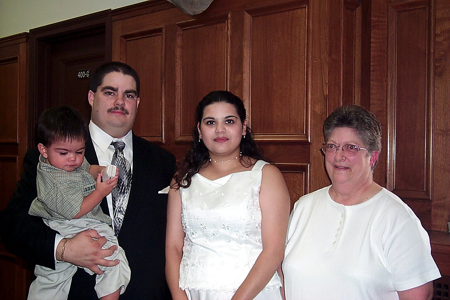 Family portrait: a man in a suit holding a young child, standing beside a woman in a white dress and an older woman in a white top, in front of wooden panels.