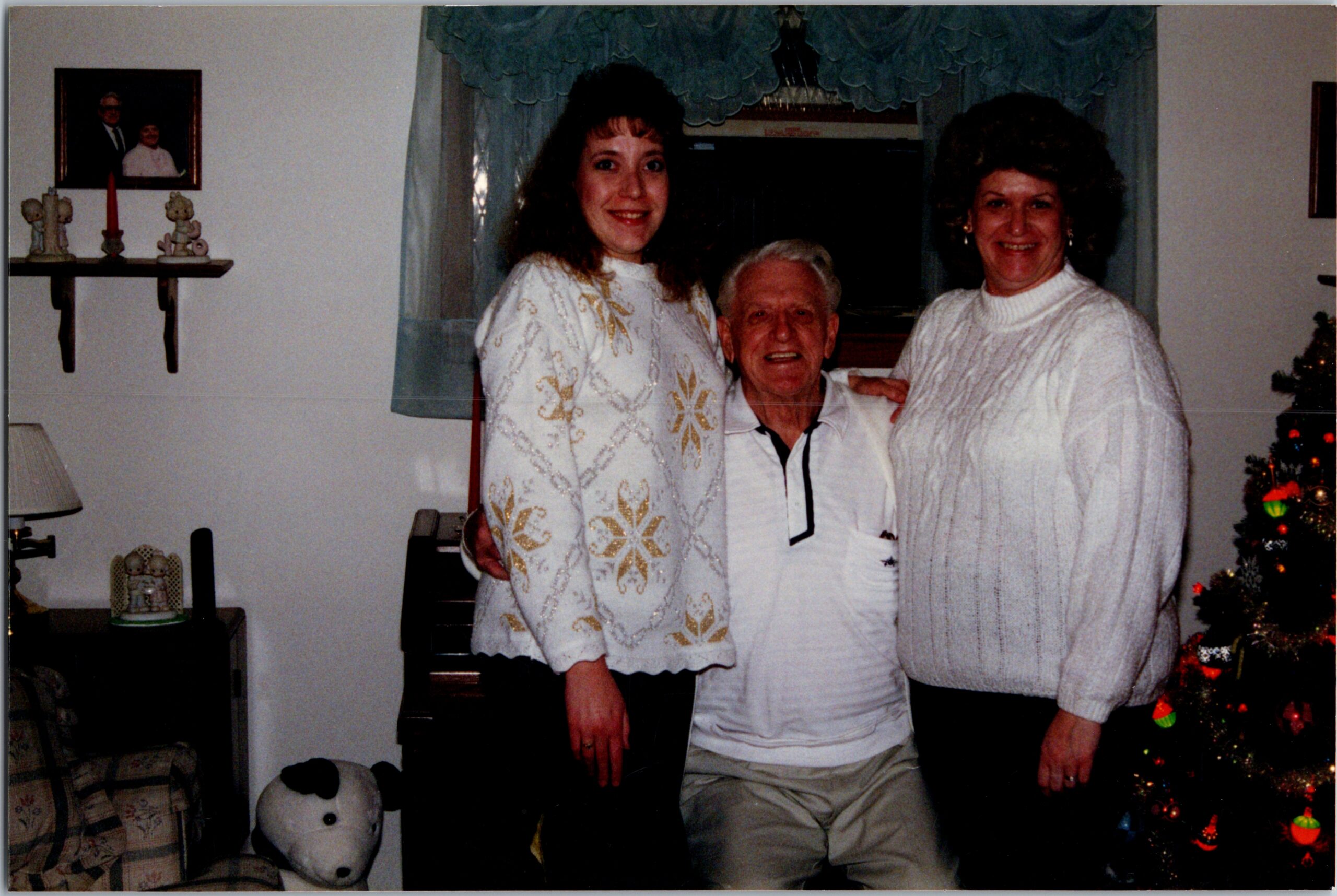 Three people posing together in a living room during Christmas; an older man sits between two women, all smiling, with a decorated Christmas tree on the right and holiday decor in the background.