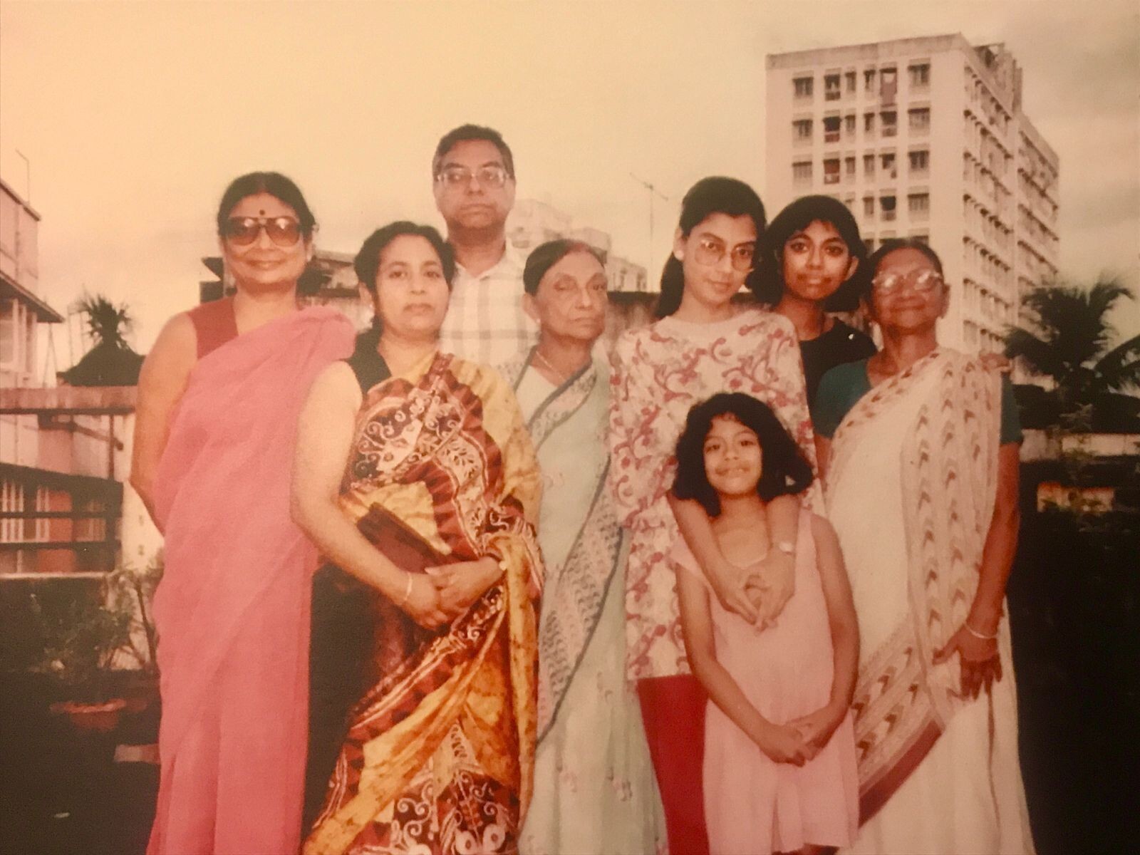 A multi-generational family poses outdoors in sarees and dresses, smiling at the camera with a tall building in the background.