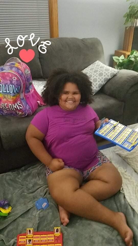 A smiling girl in a purple shirt sits on the floor playing with a blue toy xylophone, surrounded by a colorful backpack and toys on a gray rug.
