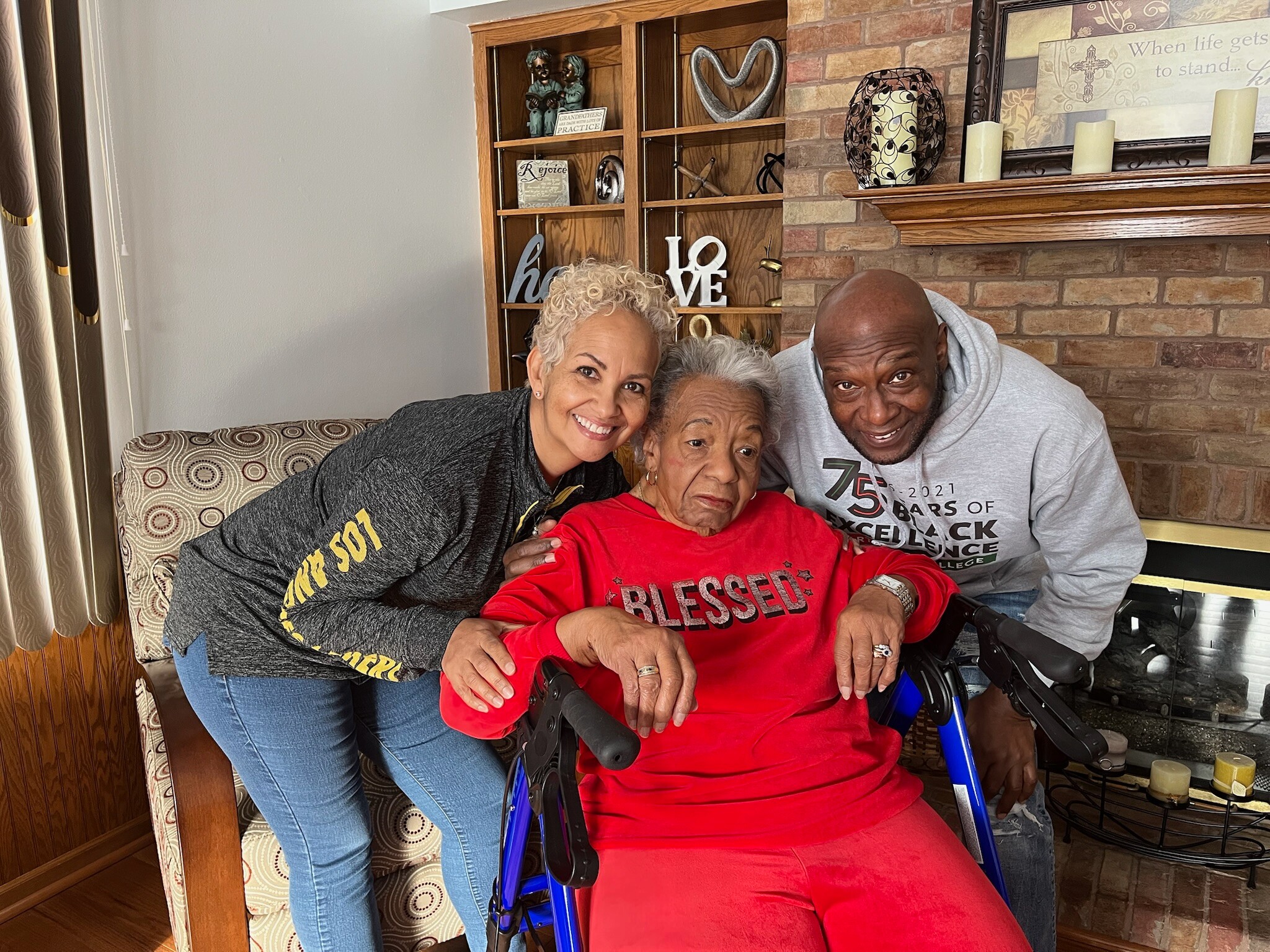 Three smiling people pose together indoors; an elderly woman in a red outfit sits in a wheelchair, flanked by two younger relatives.