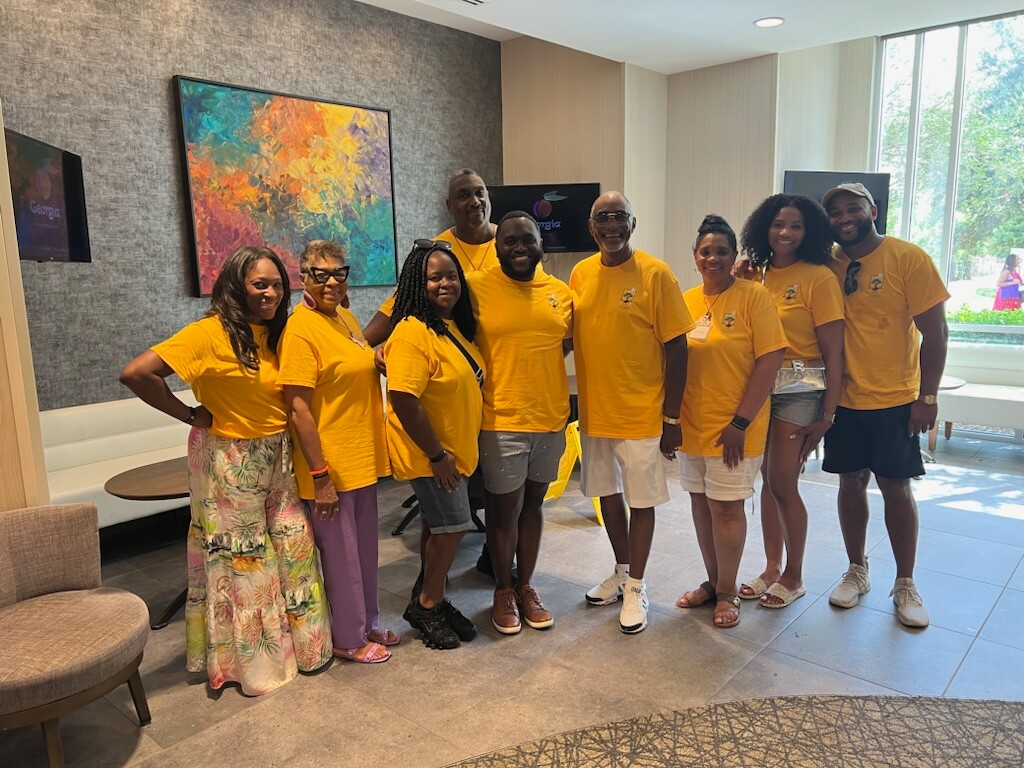 Group of nine people wearing yellow shirts posing together in a bright lounge, with a colorful abstract painting behind them.