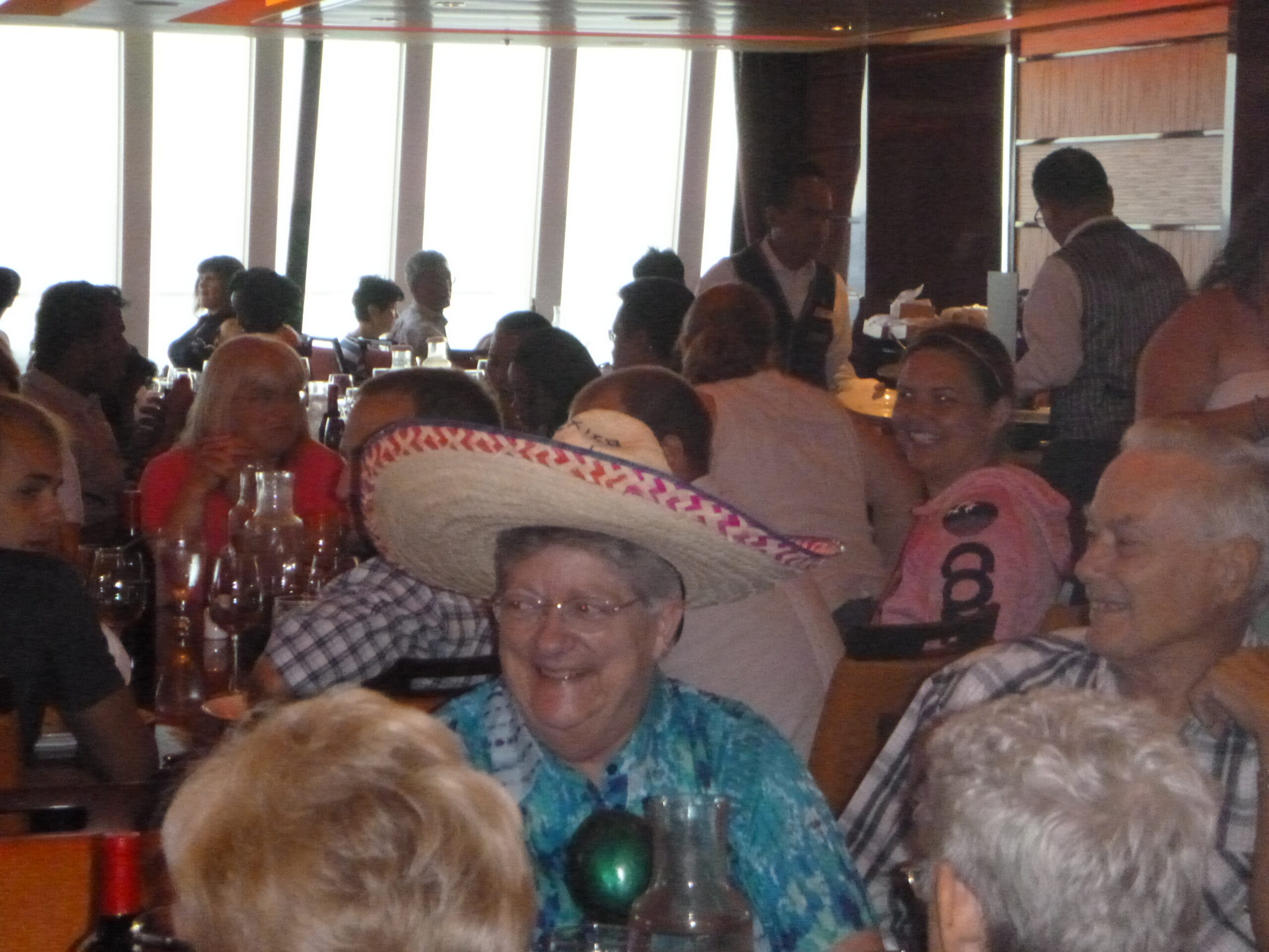 Crowded restaurant dining room with people seated at tables and a waiter in a vest in the back.