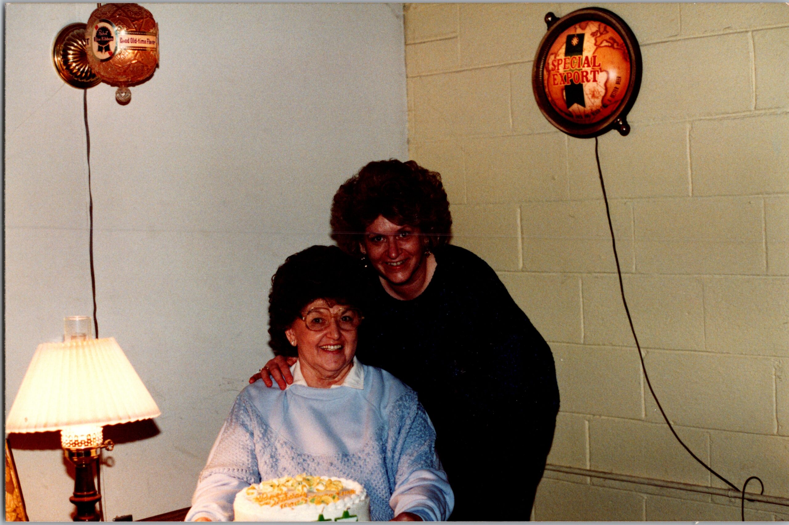 Two women at a birthday party with a cake, balloons on the wall in the background.