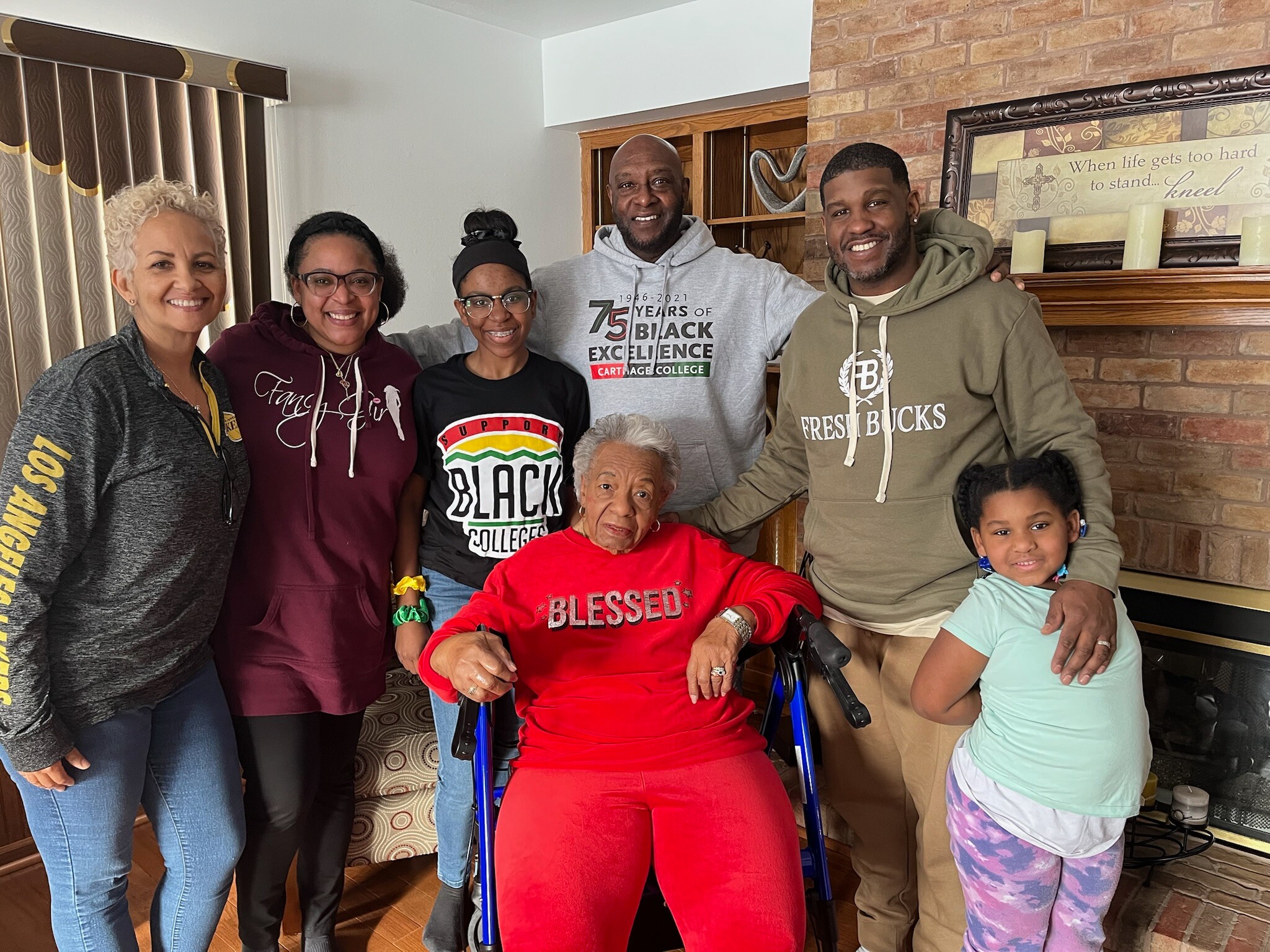 Group of six people smiling for a casual indoor photo, including an elderly woman in a red outfit in a wheelchair and a young girl on the right, with a brick fireplace in the background.