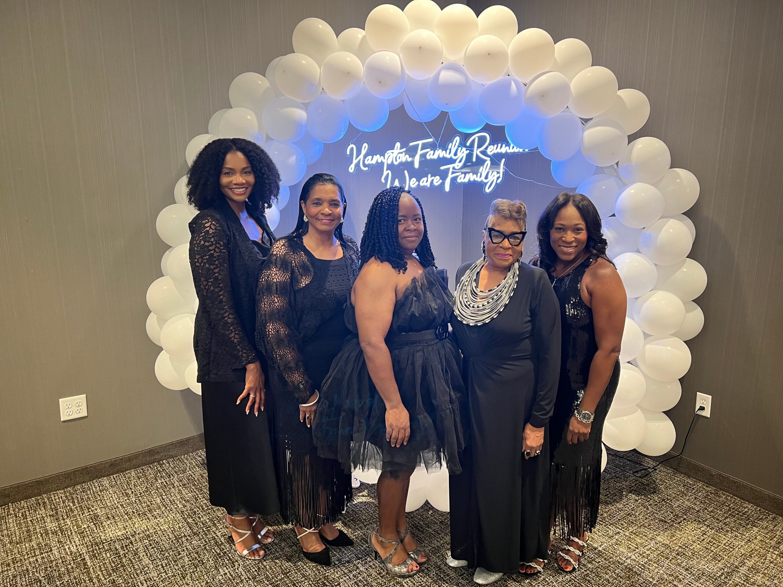 Five women in black formal dresses pose together under a white balloon arch with a neon sign reading 'Hamilton Family Reunion: We are Family'.