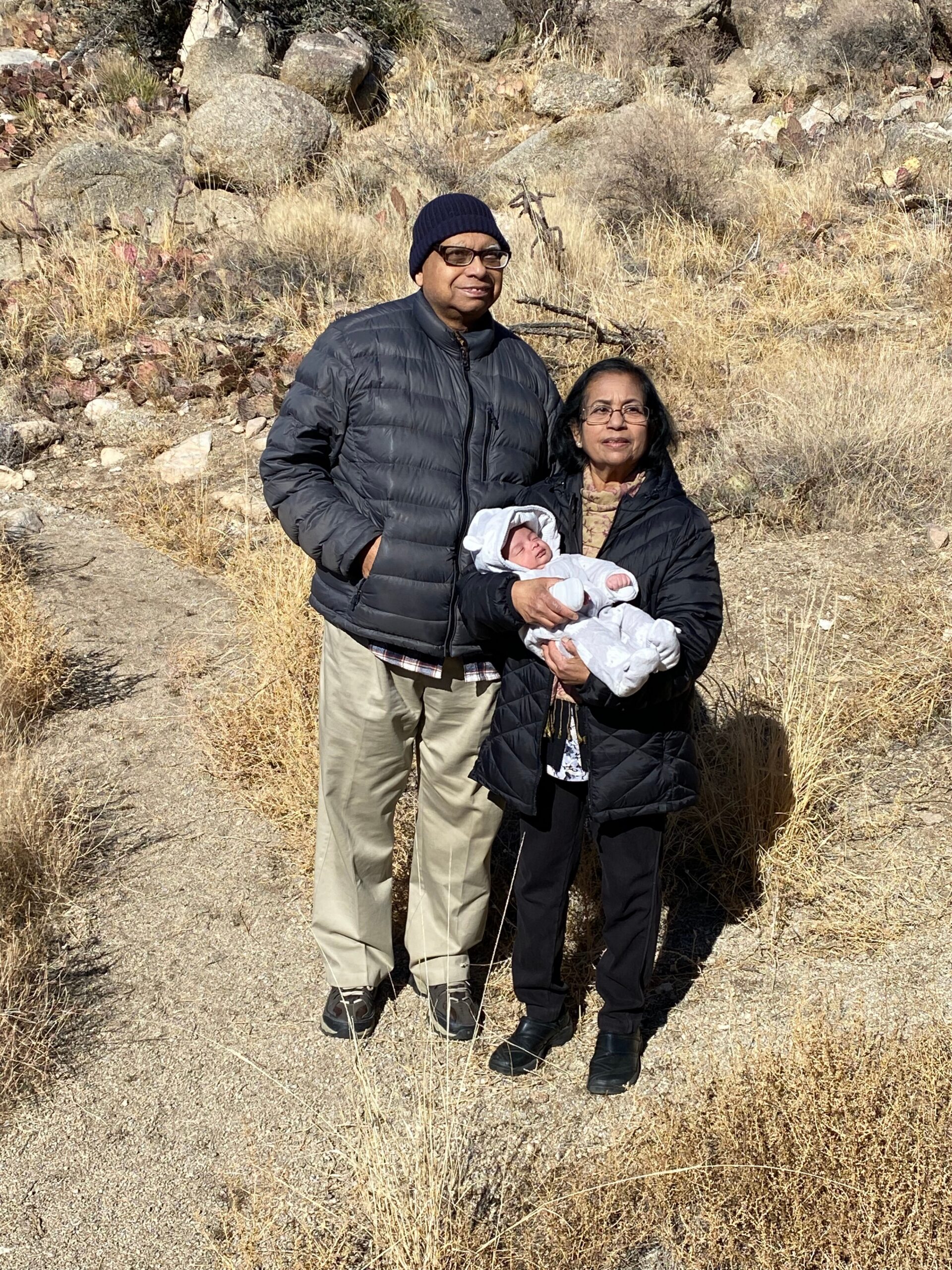 A man in a dark puffer jacket and navy beanie stands beside a woman in a black coat who holds a swaddled baby, in a dry, rocky hillside.