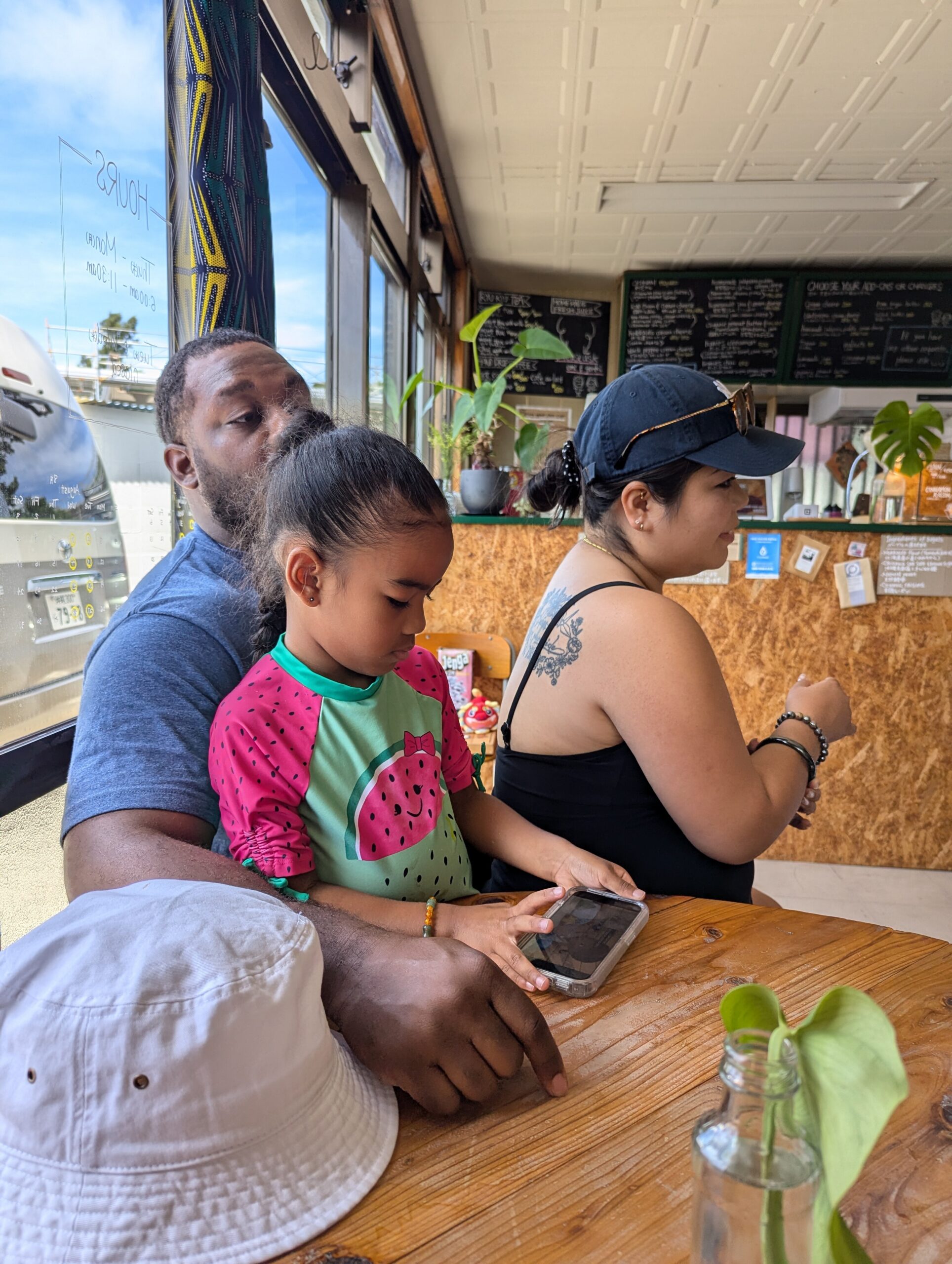 A man holds a child on his lap at a cafe counter, both looking down at a phone on a wooden table with a woman nearby.