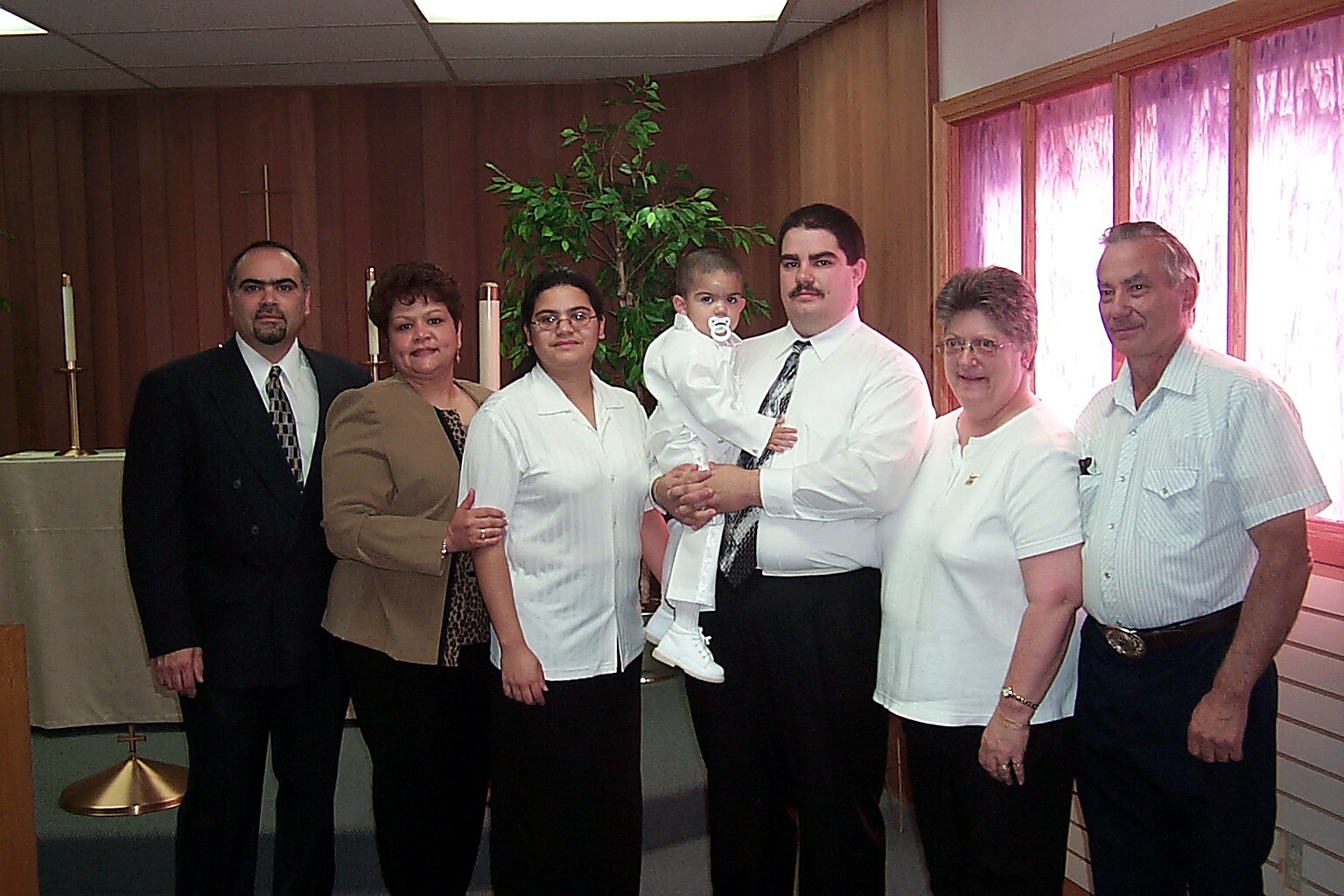 Group of seven adults and a baby posed for a photo in a church setting; man in a white shirt holds a baby while others stand beside him.