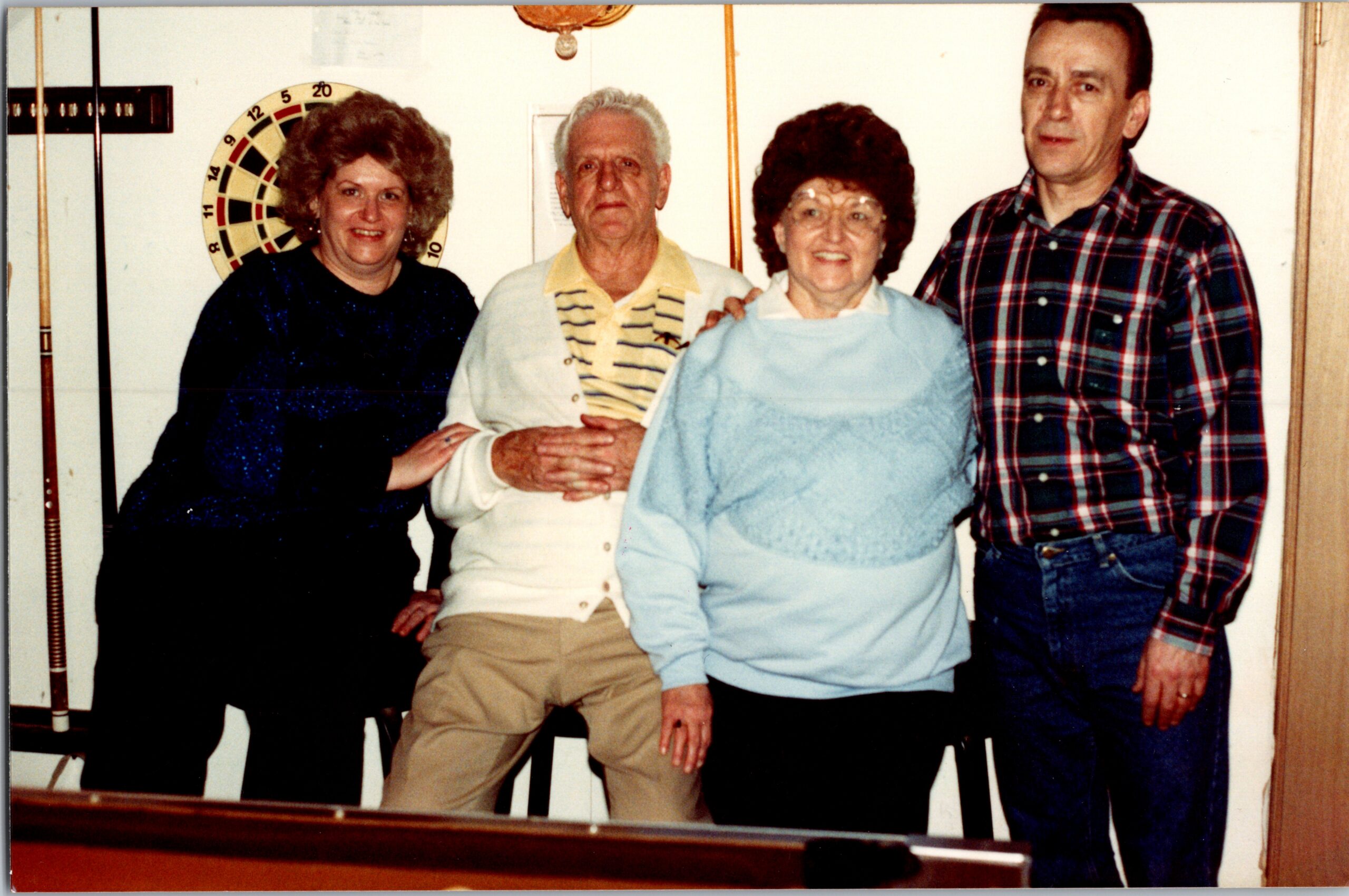 Four adults posing together indoors in a casual photo, with a dartboard on the wall behind them.