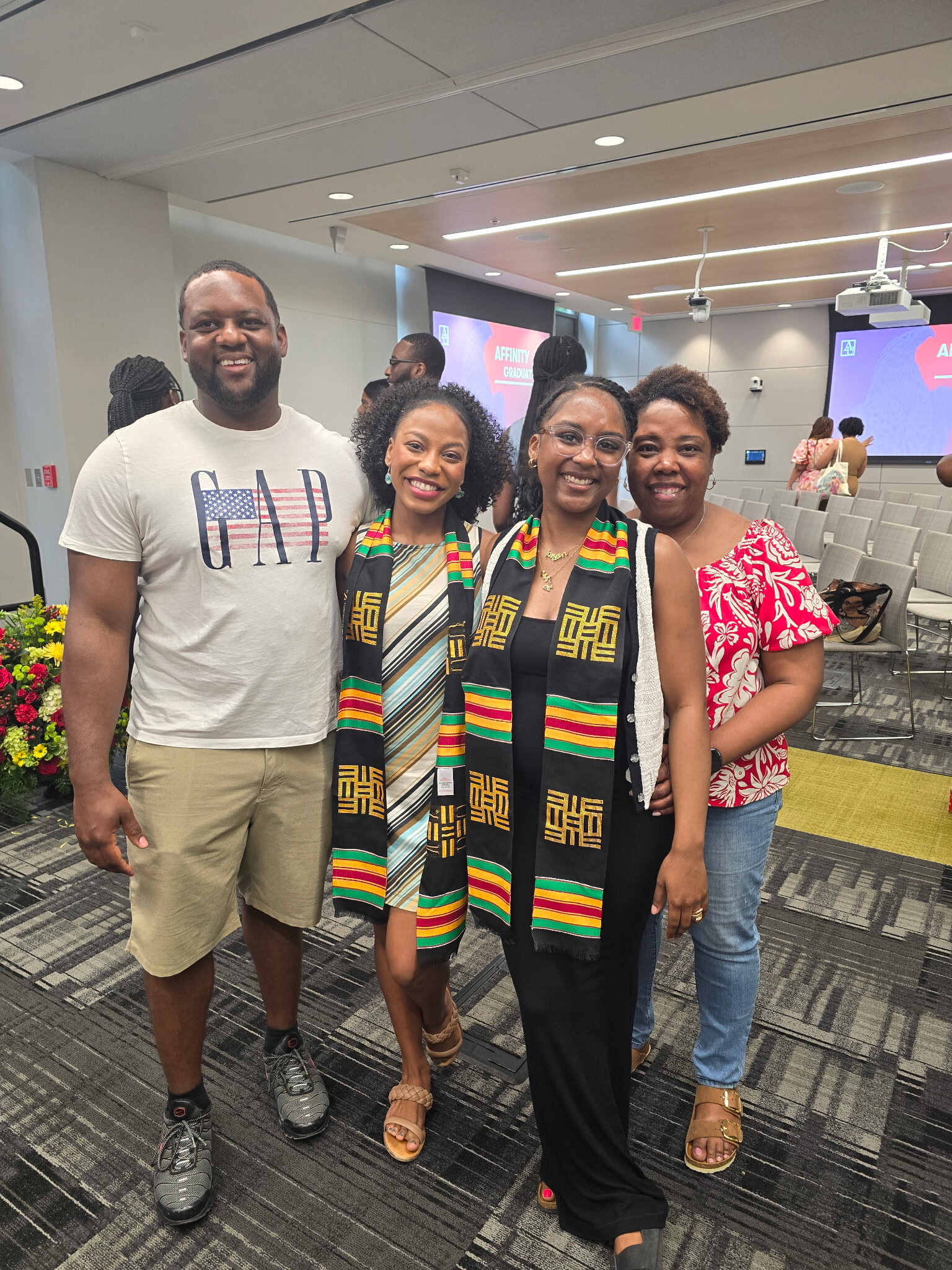 Four adults posing for a group photo in a conference room, with two women wearing colorful kente scarves and a man in a white GAP shirt on the left.