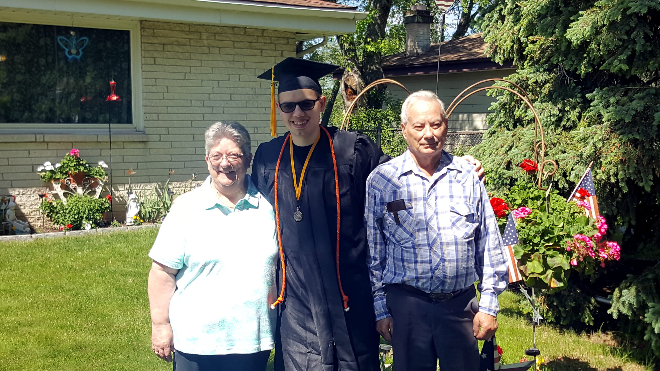 Graduate in cap and gown posing with an elderly couple in a sunny front yard celebration.