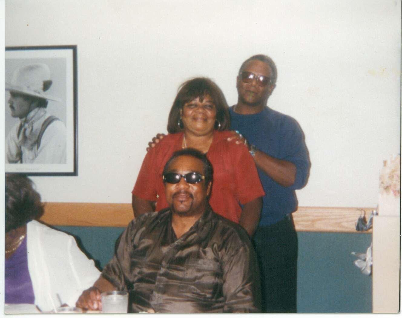 Three people posing in a casual dining setting: a man in front wearing sunglasses and a brown patterned shirt, a smiling woman in red behind him, and a man in blue standing behind her with hands on her shoulders.