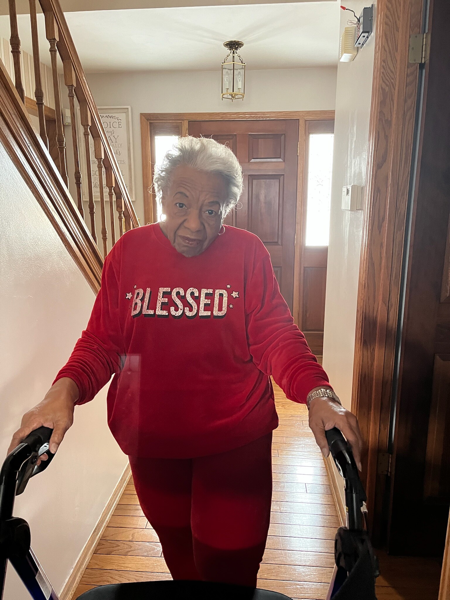 Older woman in a red sweatshirt with 'BLESSED' on the chest, using a walker in a home hallway.