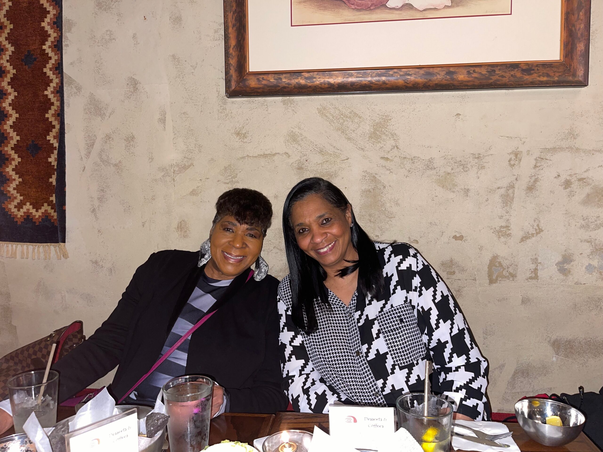 Two women sit closely at a table in a restaurant, smiling for the camera with drinks and desserts in front of them.