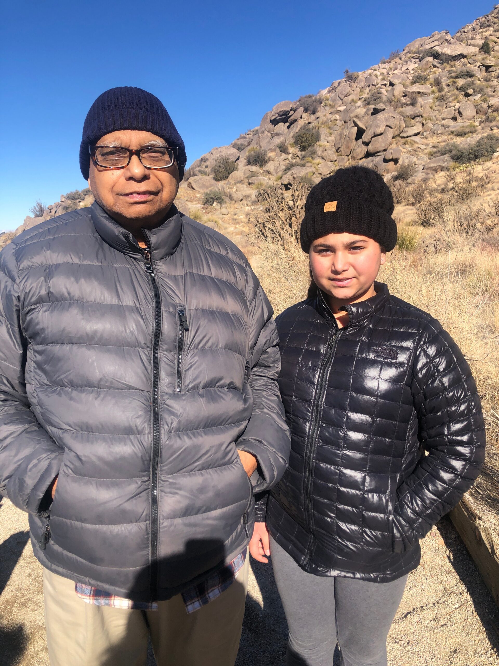 Two people wearing warm jackets stand outdoors on a rocky hillside under a clear blue sky.