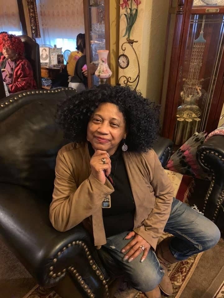 Older woman with curly hair sits on a black leather chair in a cozy living room, hands folded under her chin.