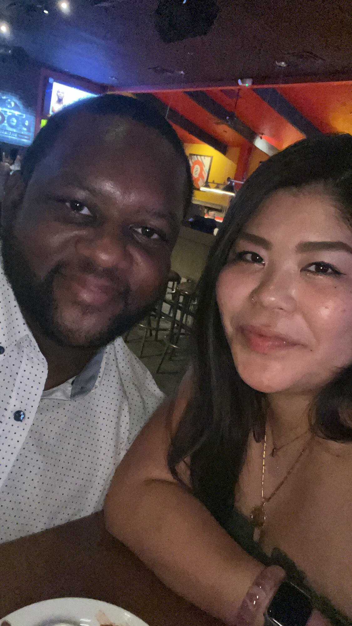 Two friends taking a close-up selfie at a dimly lit bar or restaurant, smiling with colorful decor in the background.