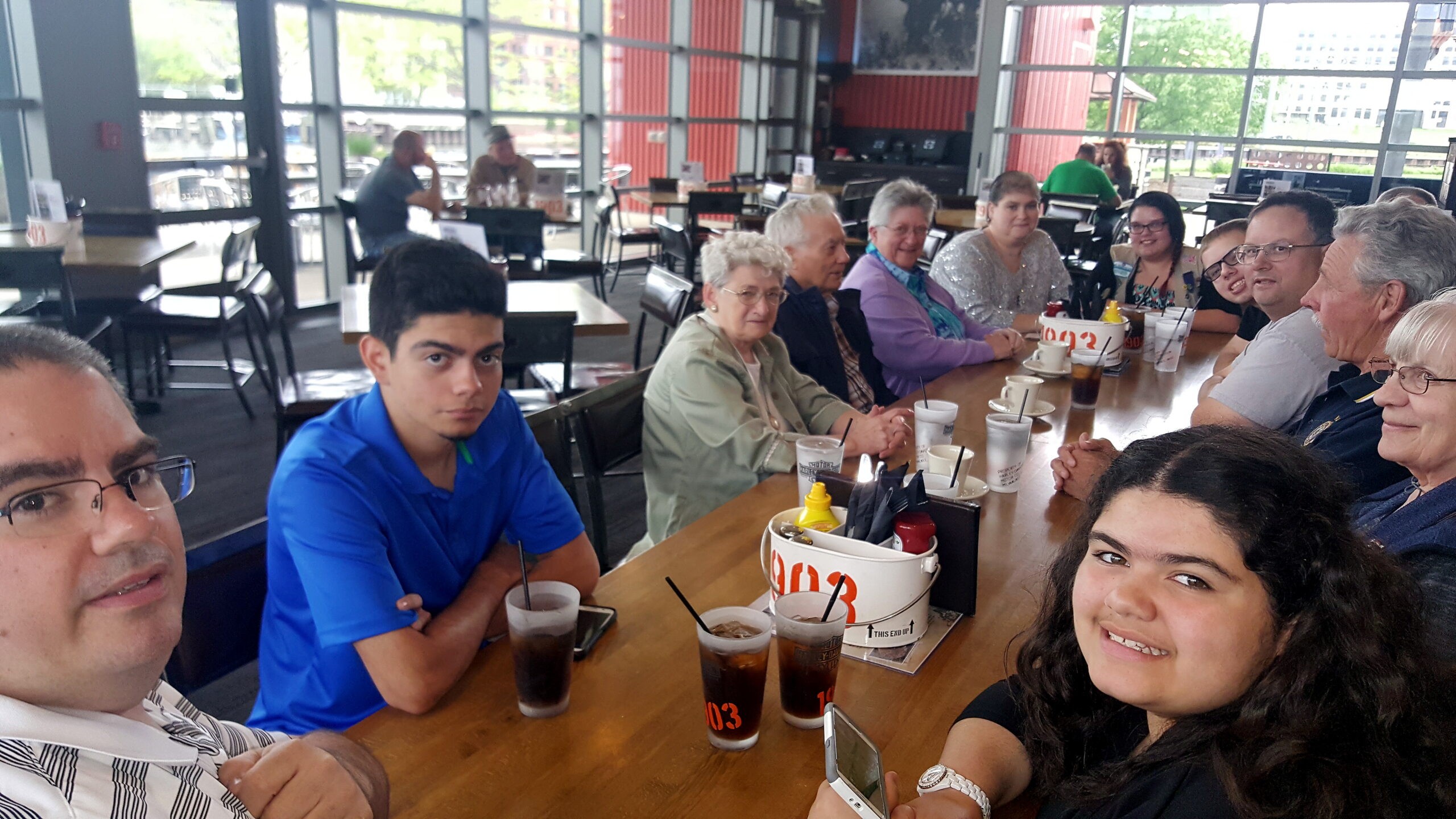 Group of adults gathered around a long wooden table in a bright casual dining space, drinks with straws on the table, taking a group selfie at the camera edge in the foreground.