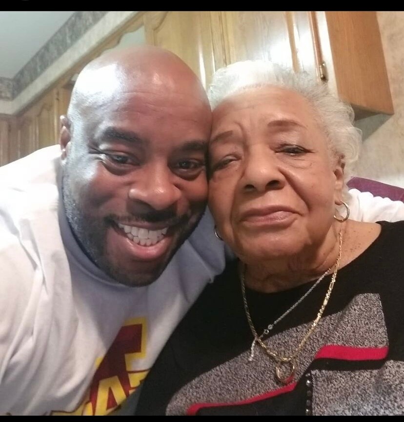 Close-up selfie of a smiling man and an older woman cuddled together in a home setting, likely a kitchen, both looking at the camera.