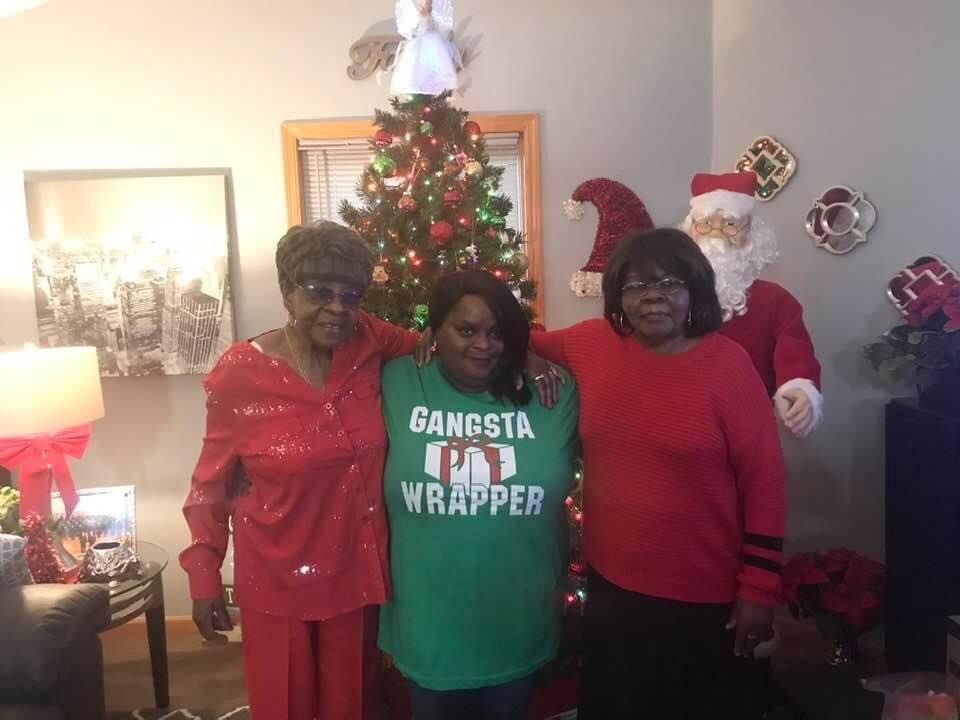 Three women pose indoors in front of a lit Christmas tree and a Santa figure in a festive living room scene.
