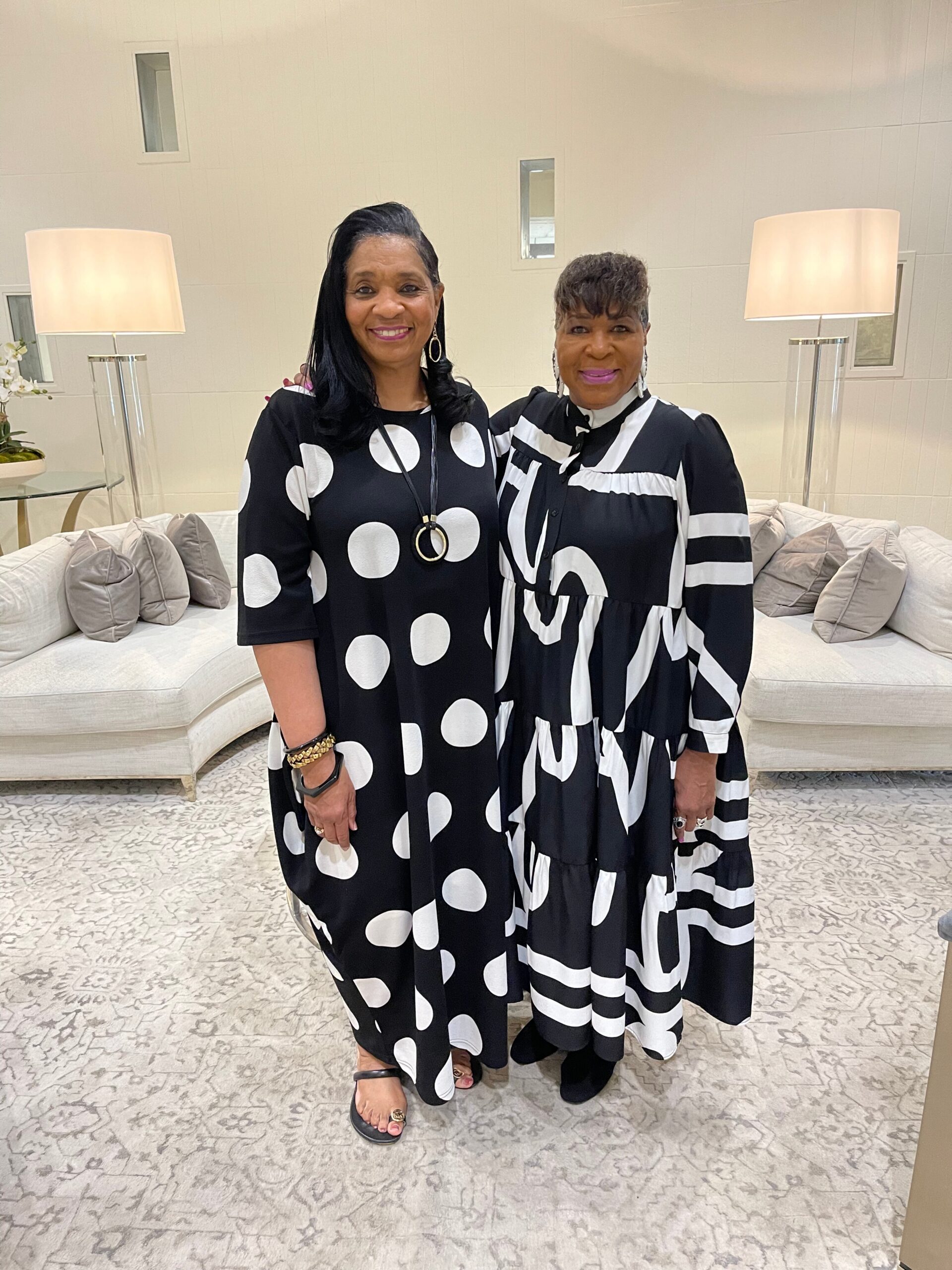 Two women in bold black-and-white polka dot outfits pose together in a bright lounge with a white sofa and neutral decor.