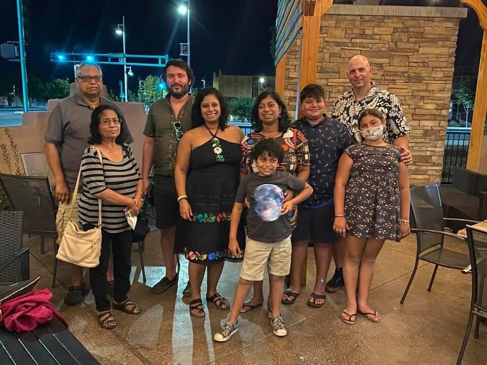 Group of nine people posing together outdoors at night near a stone column and patio furniture; everyone smiling or relaxed, some wearing casual outfits and one child in front.