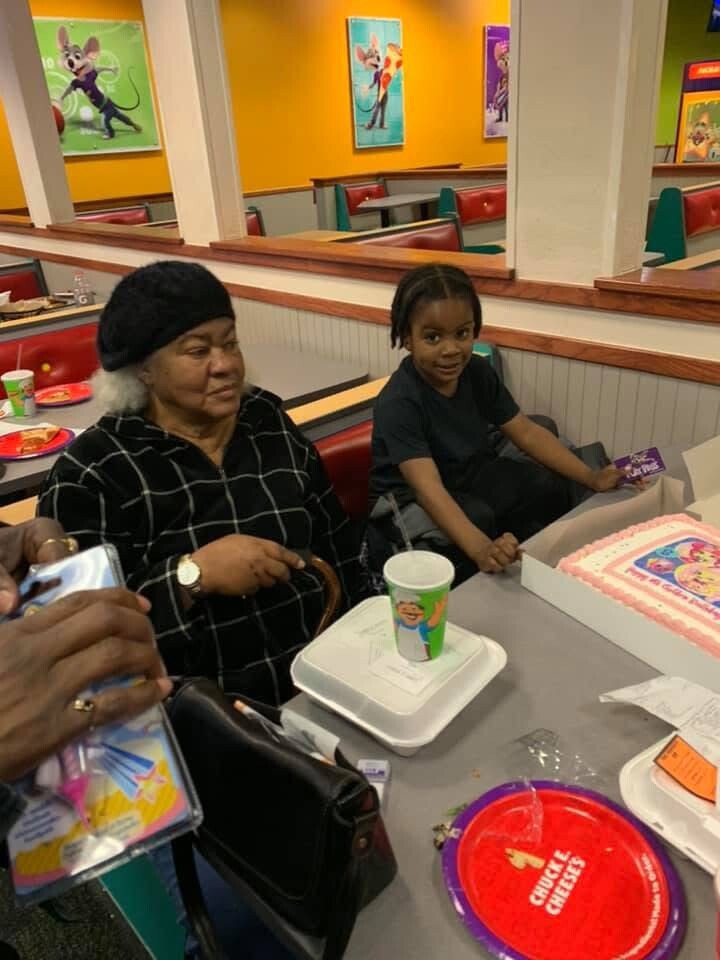 A child and an older woman sit at a restaurant table with a pink birthday cake and party supplies nearby.