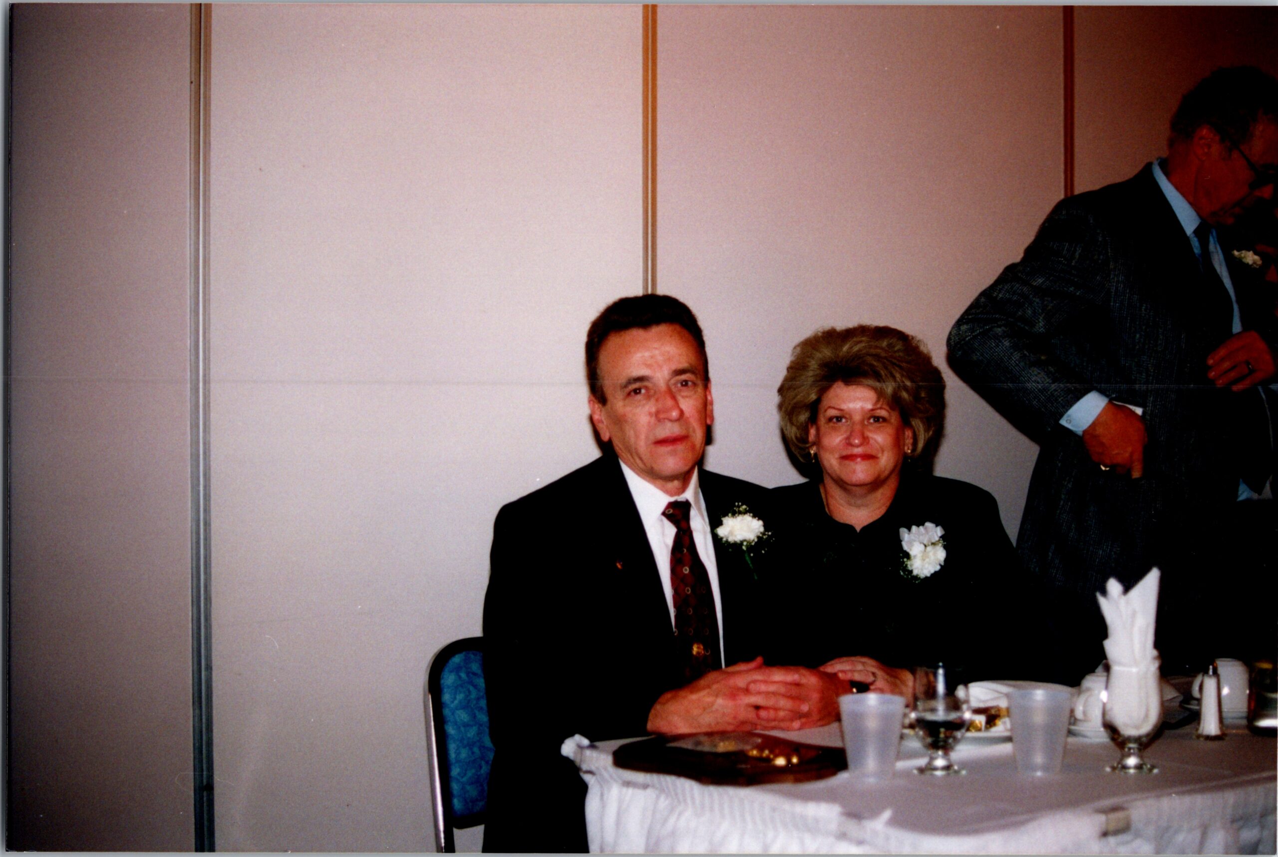 Older couple in formal attire seated at a wedding reception table, with a standing man beside them. They have boutonnieres and are dining with plates and drinks in front of them.