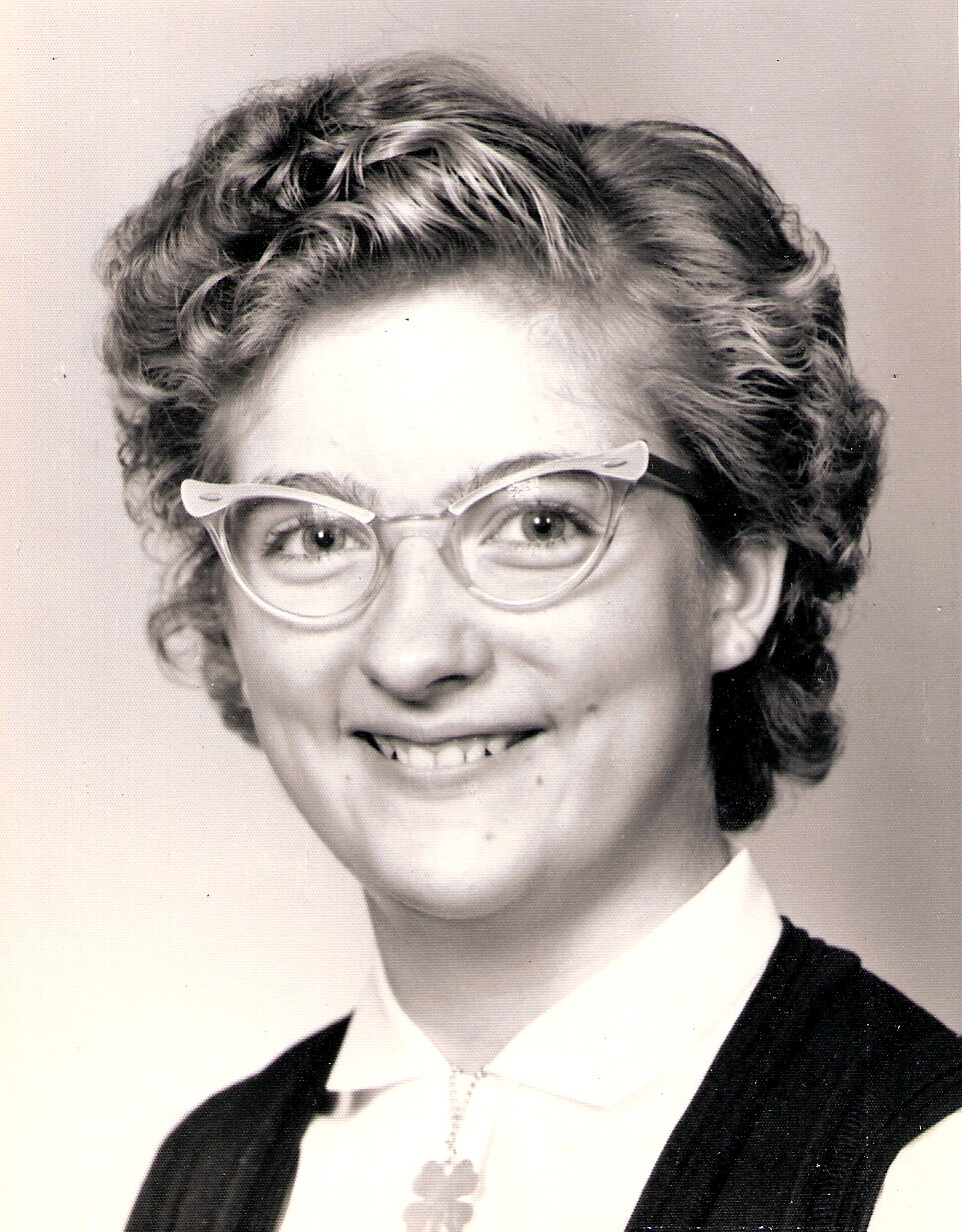 Smiling woman with short, curly hair and vintage cat-eye glasses, wearing a white collared blouse and a clover pendant necklace in a black vest.