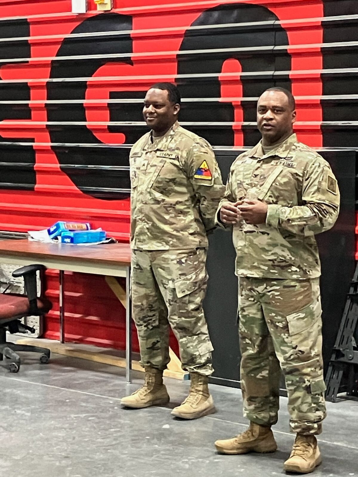 Two soldiers in camouflage uniforms stand indoors in front of a red-and-black mural, facing the camera with neutral expressions.