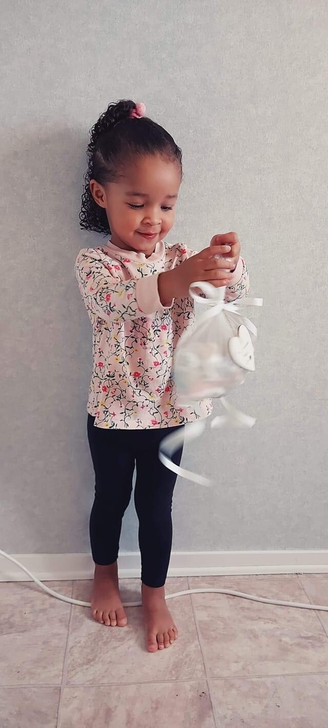 Little girl in a floral sweater and black leggings holds a white gift bag with a bow.