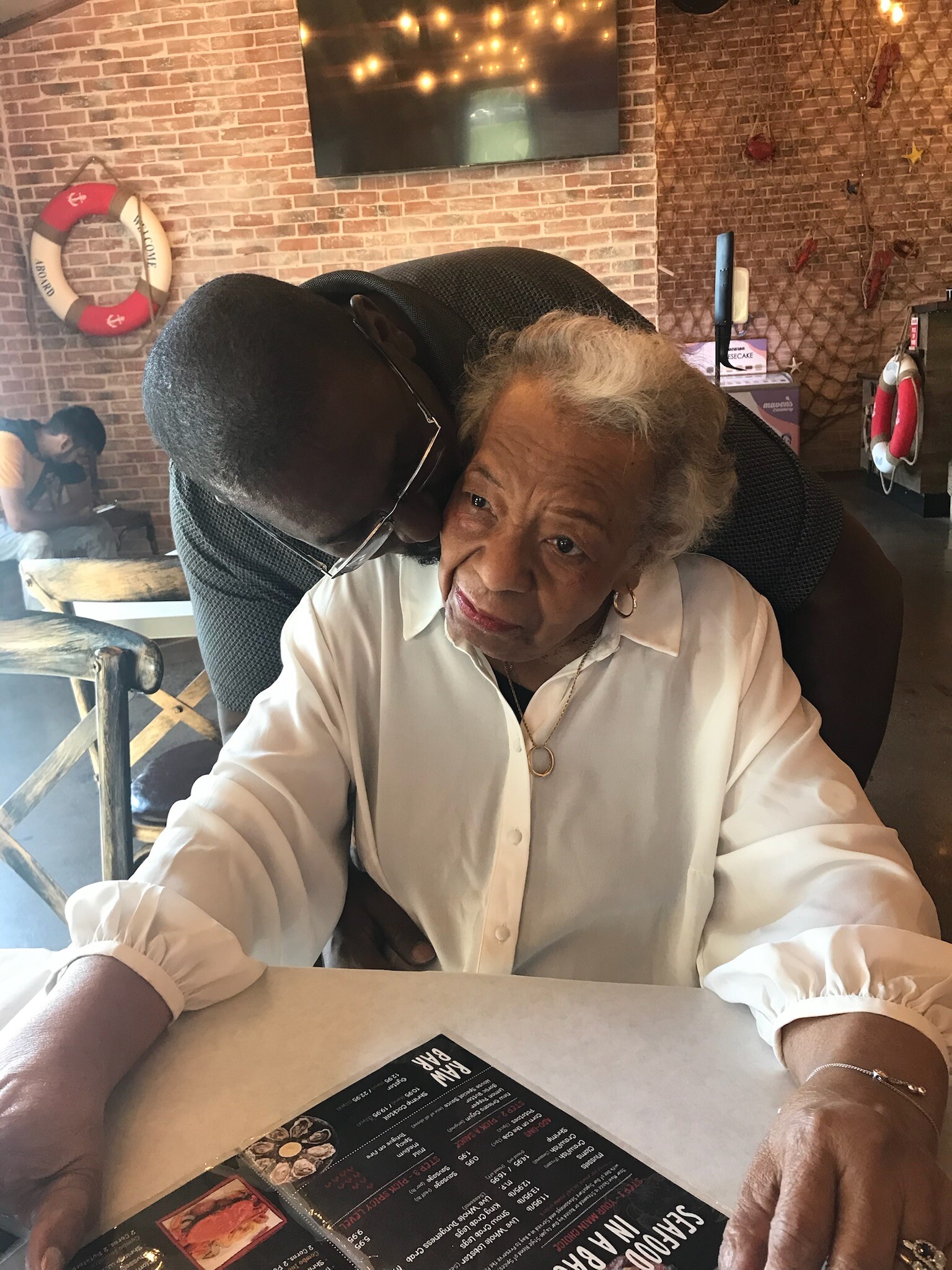 Elderly woman in a white blouse sits at a restaurant table as a man hugs her from behind, with a menu visible on the table in the foreground.