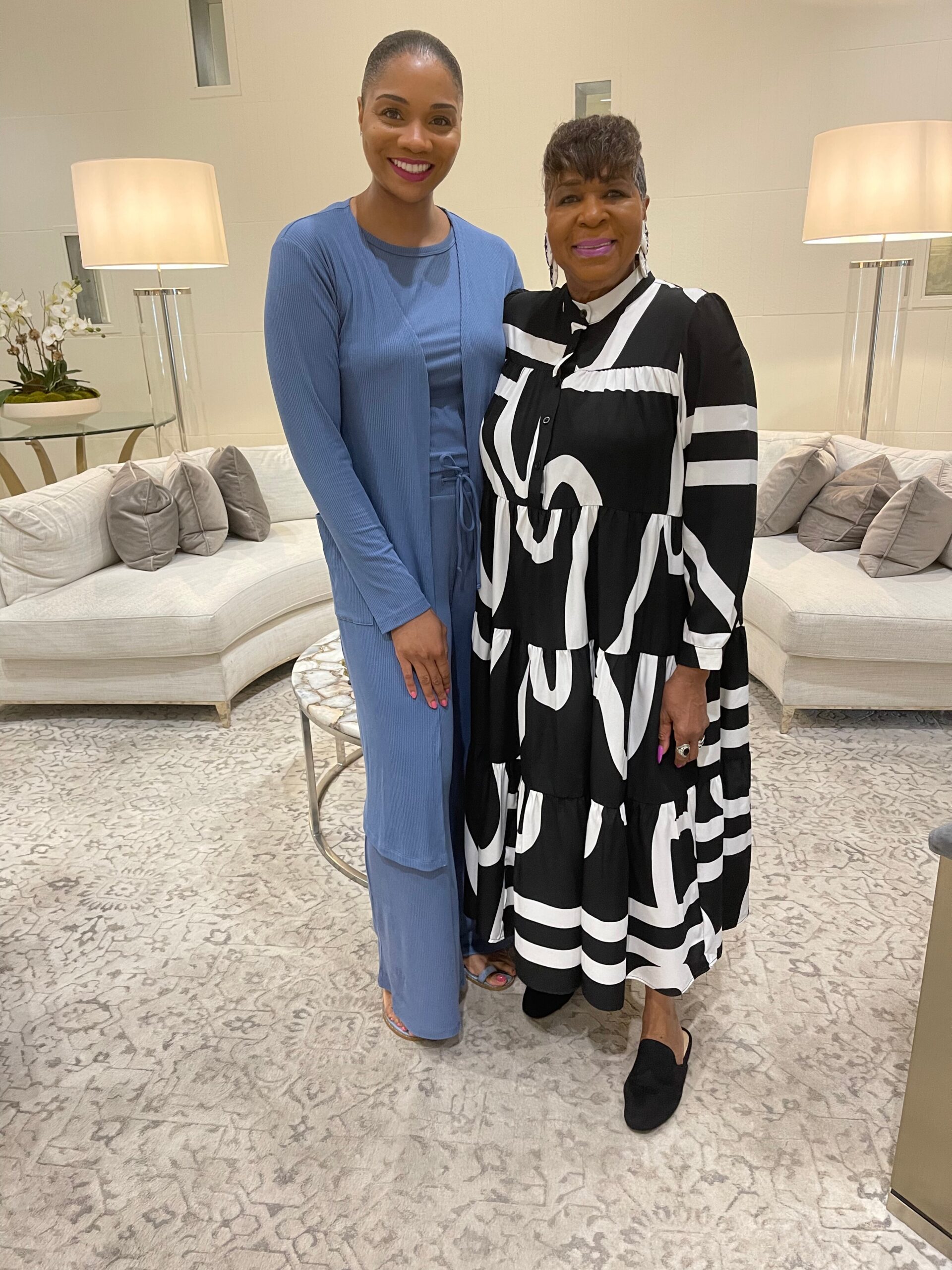 Two women posing together in a bright living room; one in a blue matching outfit, the other in a black-and-white patterned dress.