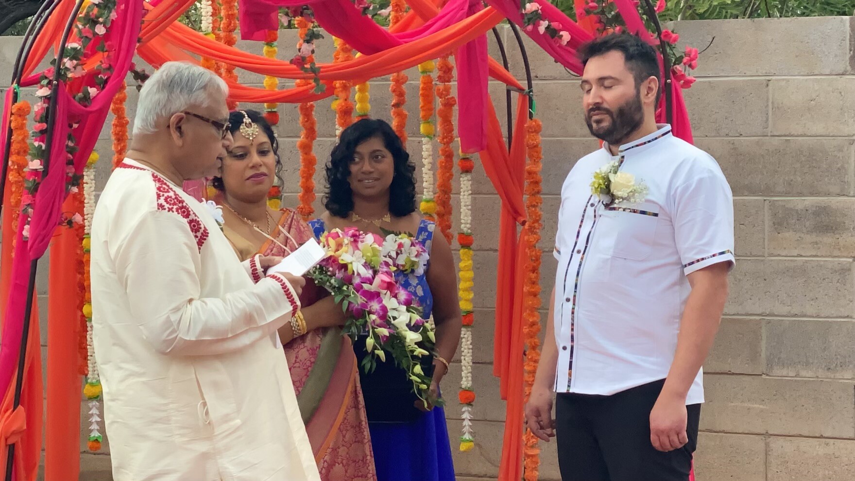A wedding ceremony with an officiant and couple under orange-pink fabric and flower garlands; the groom stands on the right, couple center-left holding bouquets.