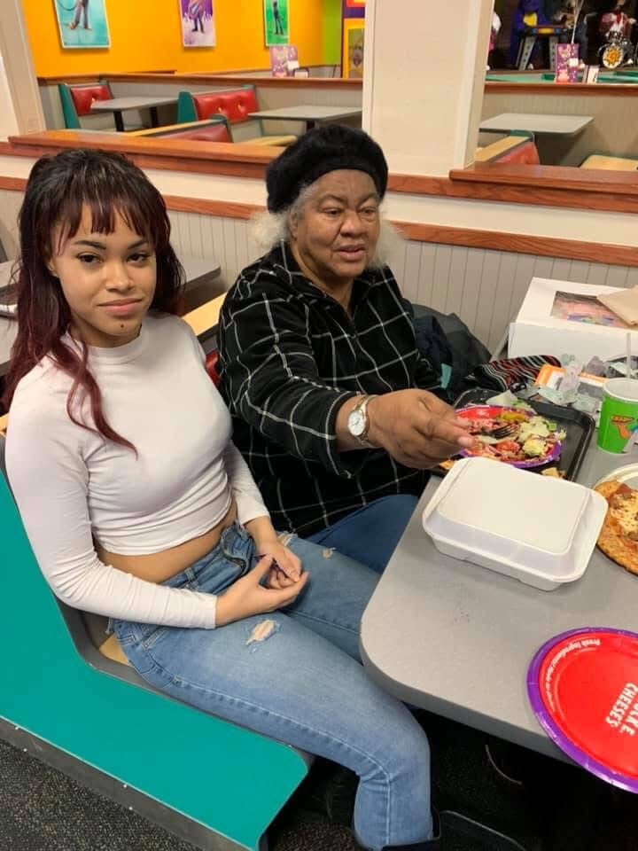 Two women sit together at a colorful fast-food booth; a young woman in a white crop top and ripped jeans beside an older woman in a black plaid shirt and hat, with food on the table.