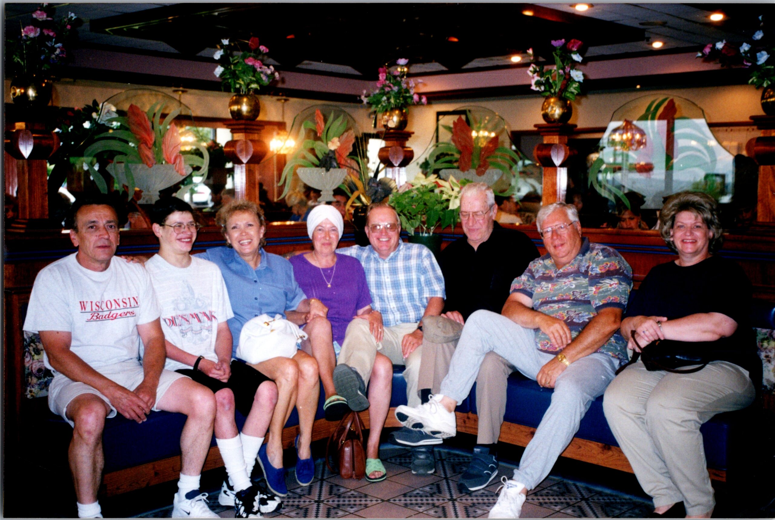Eight adults seated closely in a restaurant booth, smiling for the camera with floral decor and warm lighting behind them.