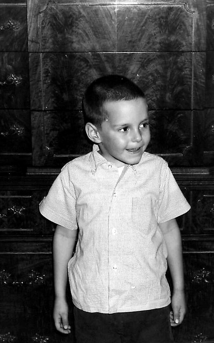 A young boy with short hair wearing a striped button-up shirt, standing and smiling to the side in front of a dark, glossy background.