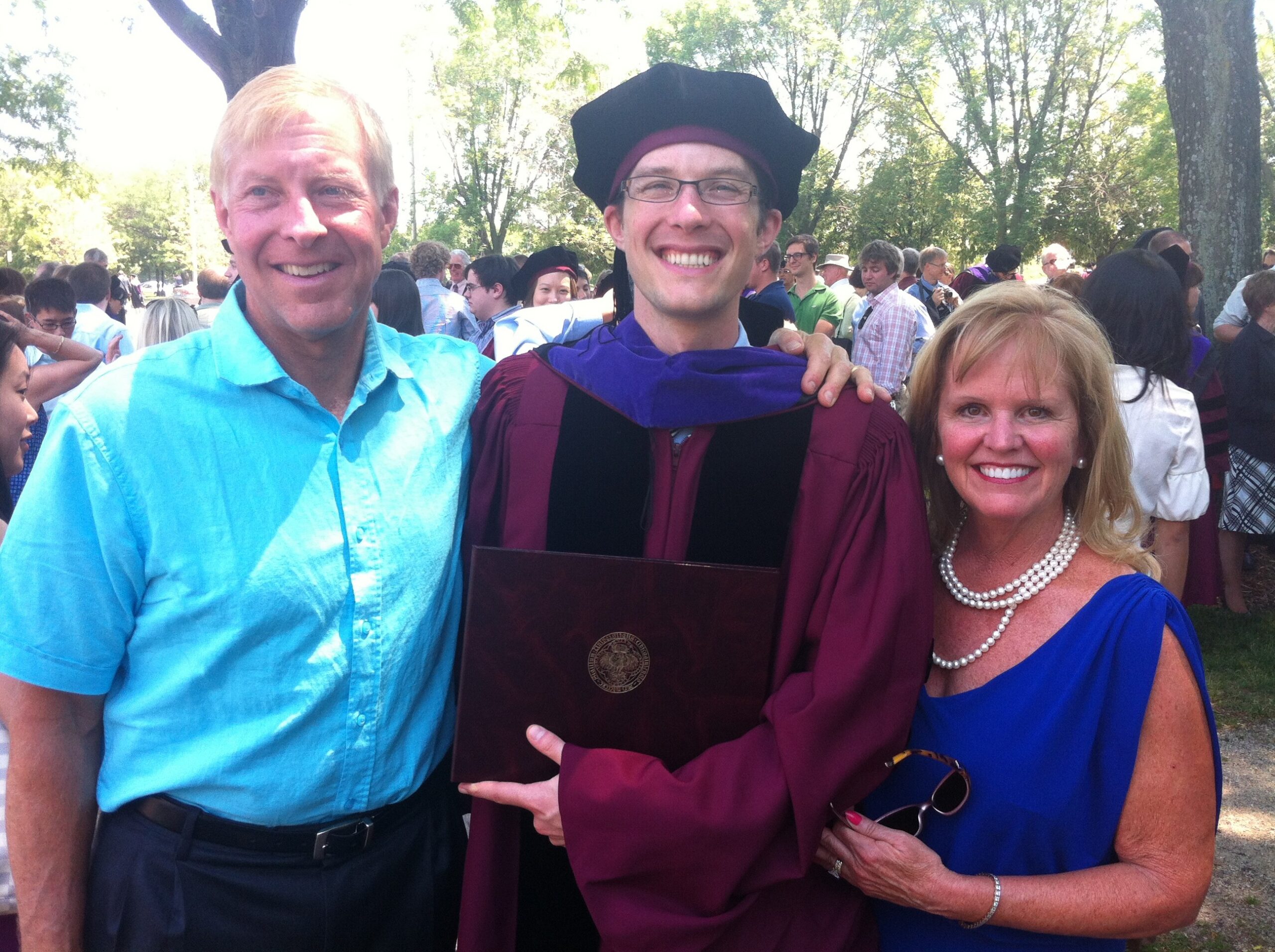 Graduate in maroon gown and cap smiles, holding a diploma outdoors with a friend/family member beside him.